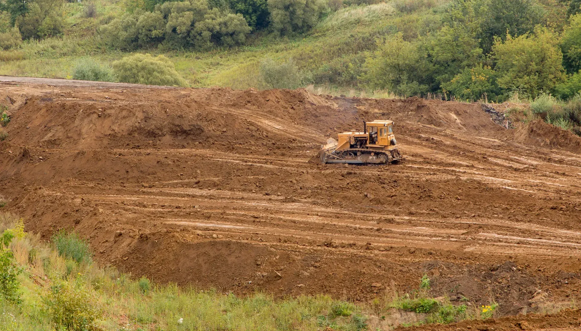 Bulldozer working on a muddy construction site with green trees in the background