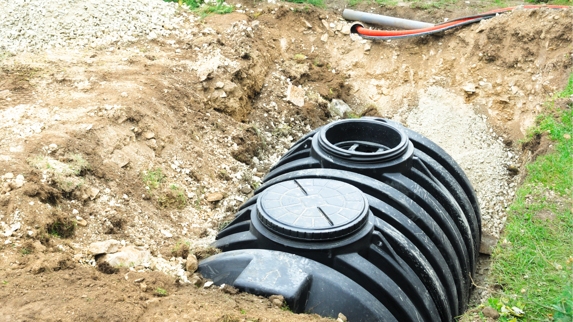 Black plastic underground tank partially buried in a dirt trench beside grass, with an open circular hatch.