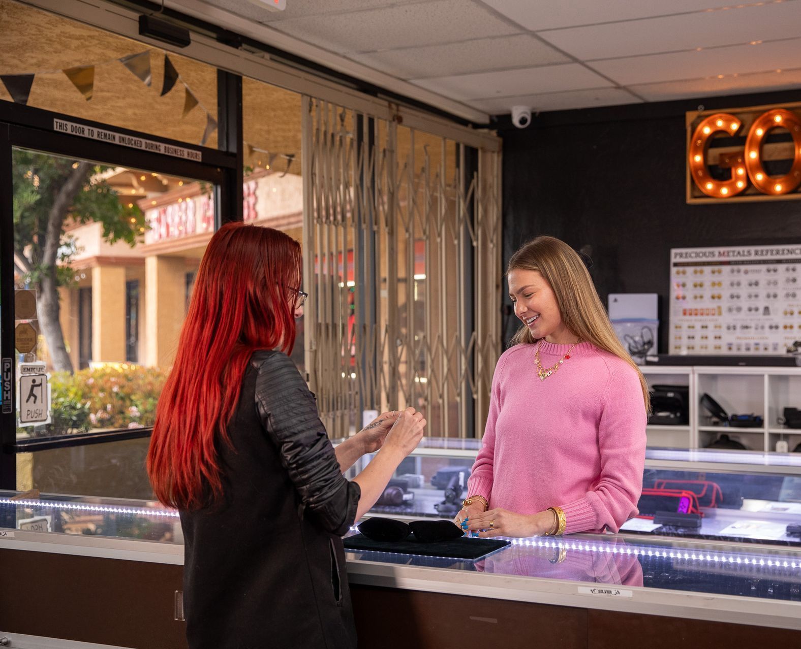 Two women talking at a jewelry store counter, one in a pink sweater and one with red hair.