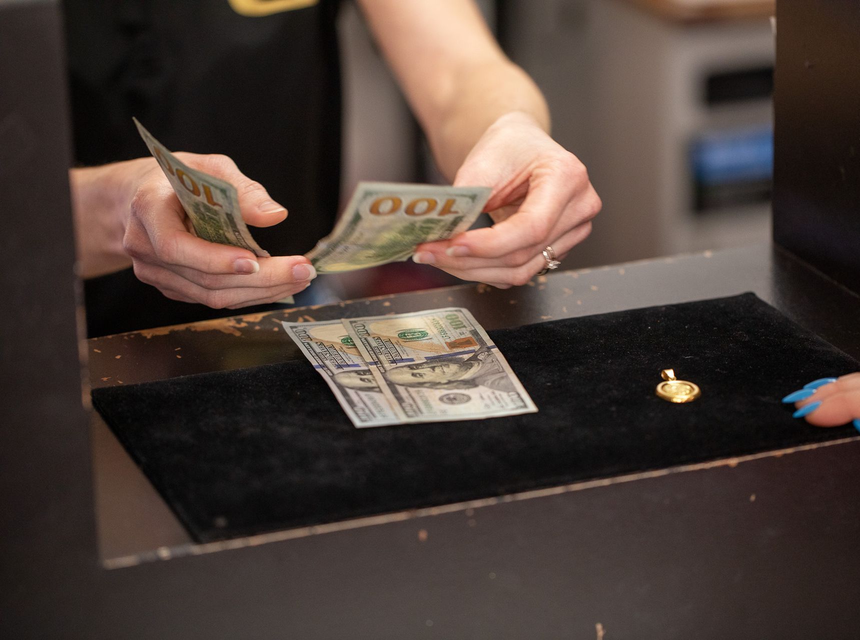 Hands counting money over a black surface, a small gold pendant lies beside it.