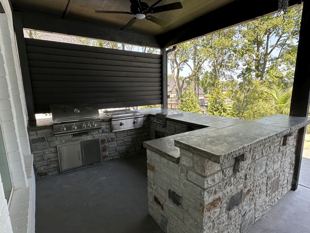 Covered outdoor kitchen with stone counters, built-in grill, and a dark wood ceiling overlooking trees