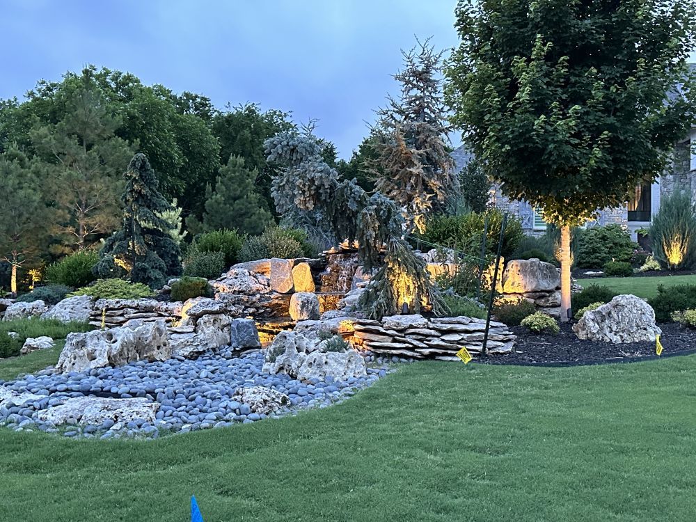 Landscaped garden with rocks, small waterfall, and trees beside a green lawn