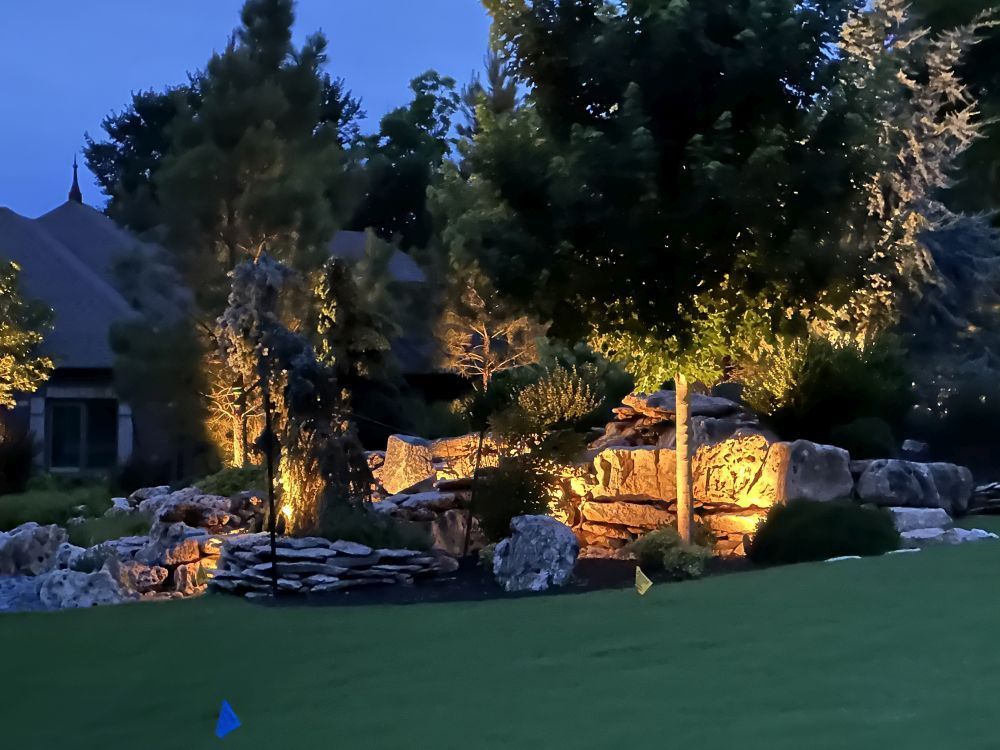 Illuminated landscaped garden at dusk with trees, rocks, and a house in the background