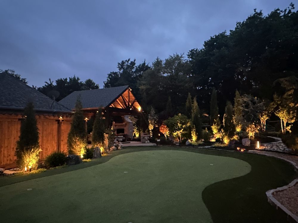 Backyard golf putting green at dusk, lit by warm landscape lights beside a house and trees