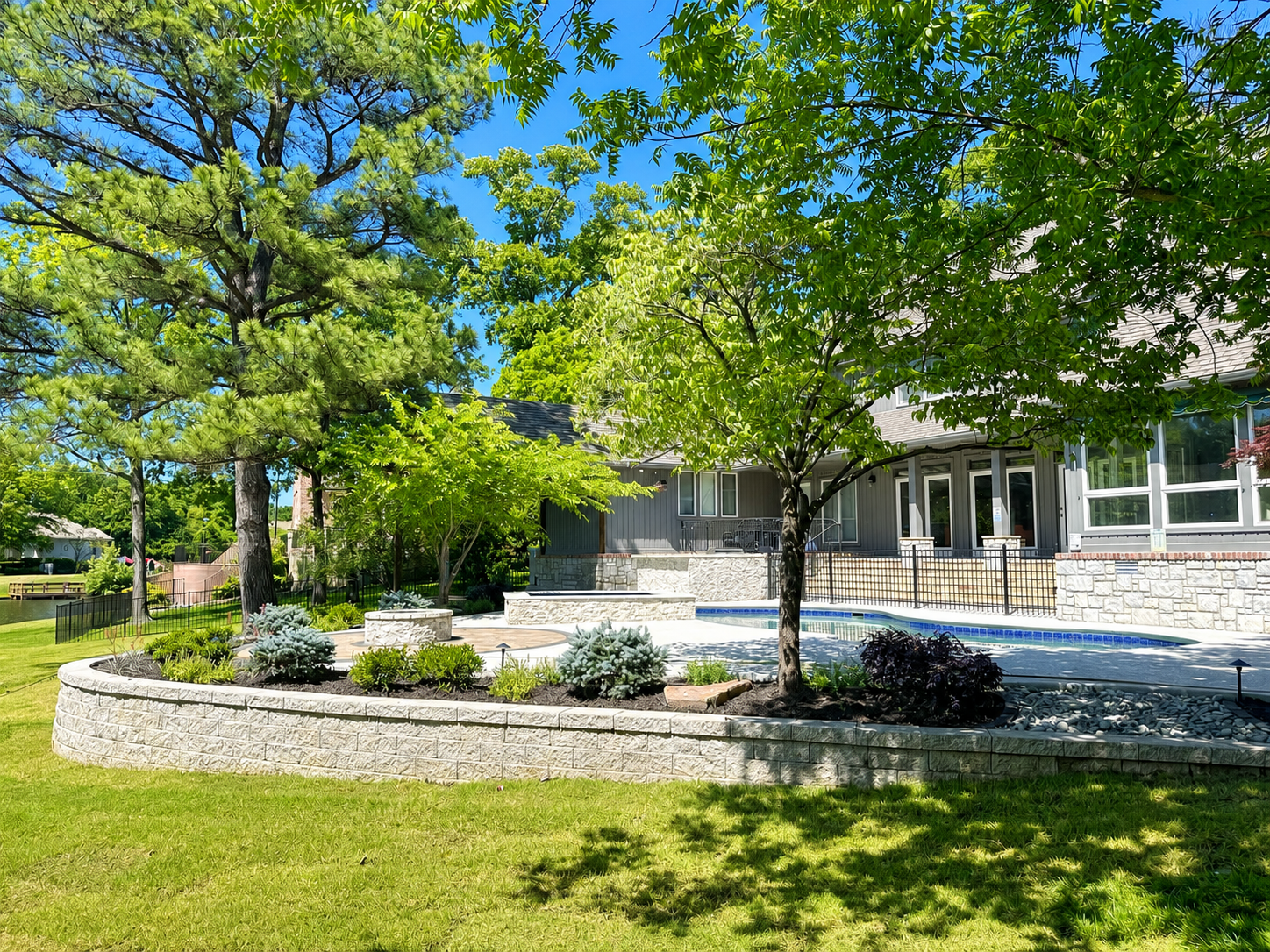 Stone retaining wall beside a paved road, with grassy field and trees under a clear blue sky.