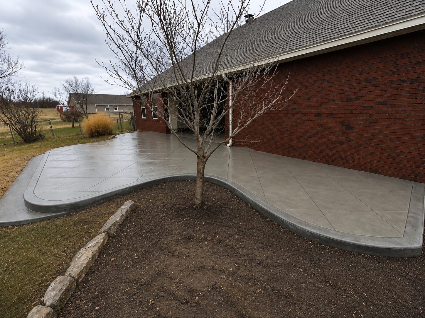Curved concrete walkway beside a brick building with young trees and a landscaped bed under cloudy skies