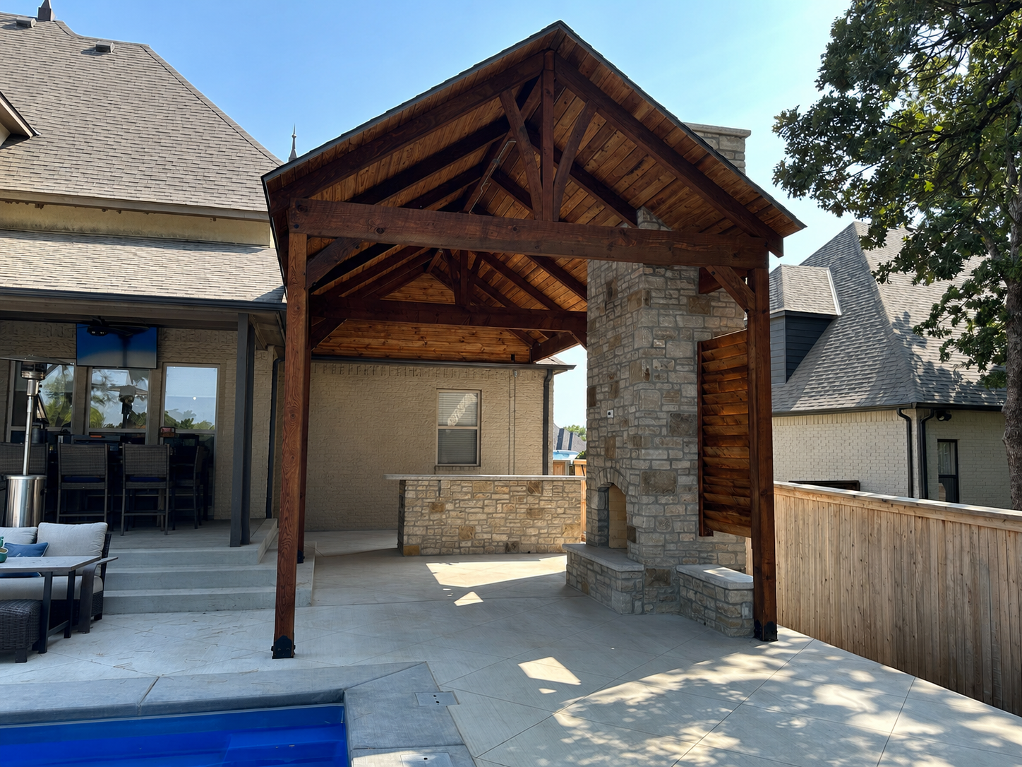 Wooden pergola beside a pool with outdoor seating and lounge chairs on a sunny patio.