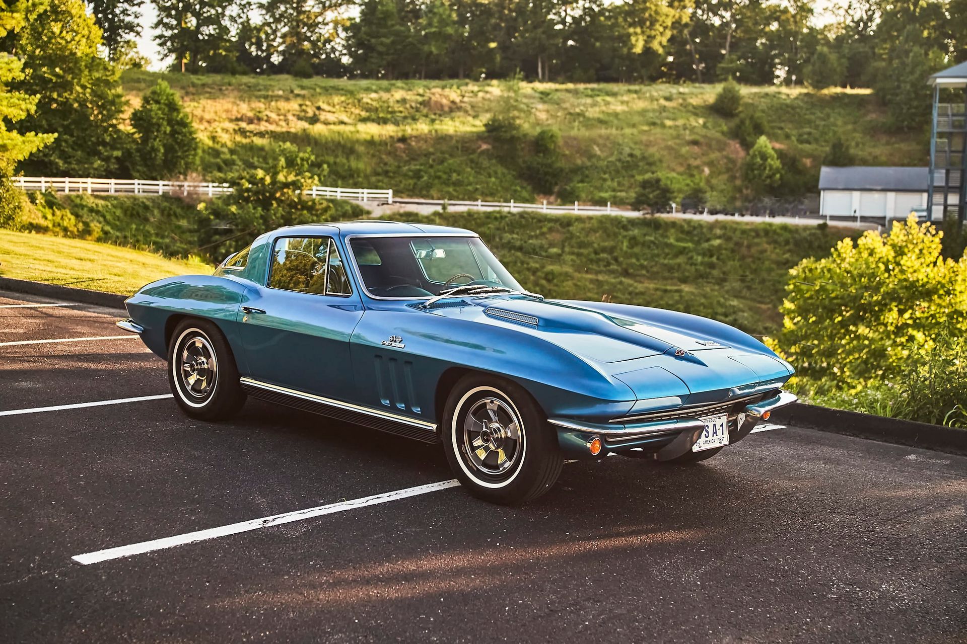 Blue vintage Corvette parked in a lot; grassy hill and building in the background.