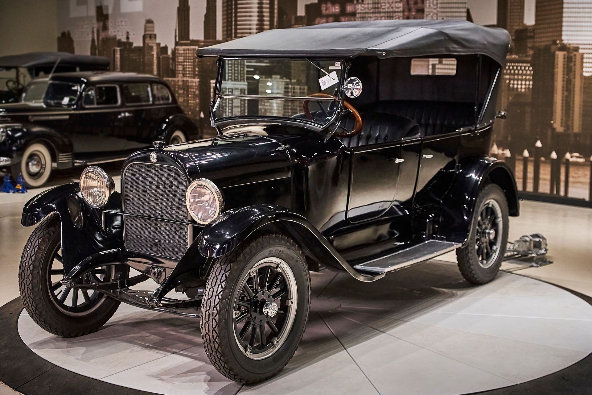 An old black car is parked in a garage with a city in the background.