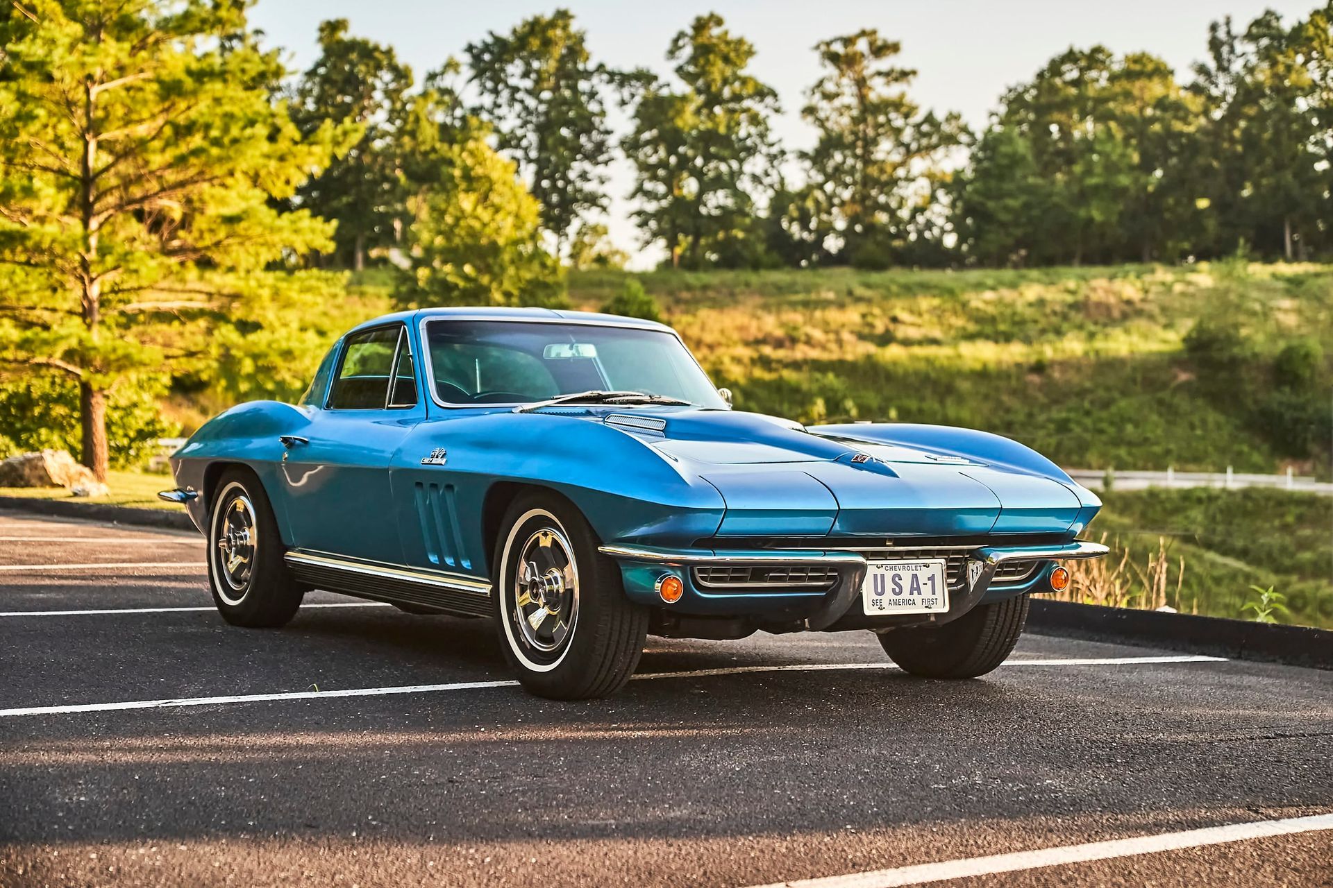 Blue classic Corvette parked on asphalt in front of green trees and a grassy hill.