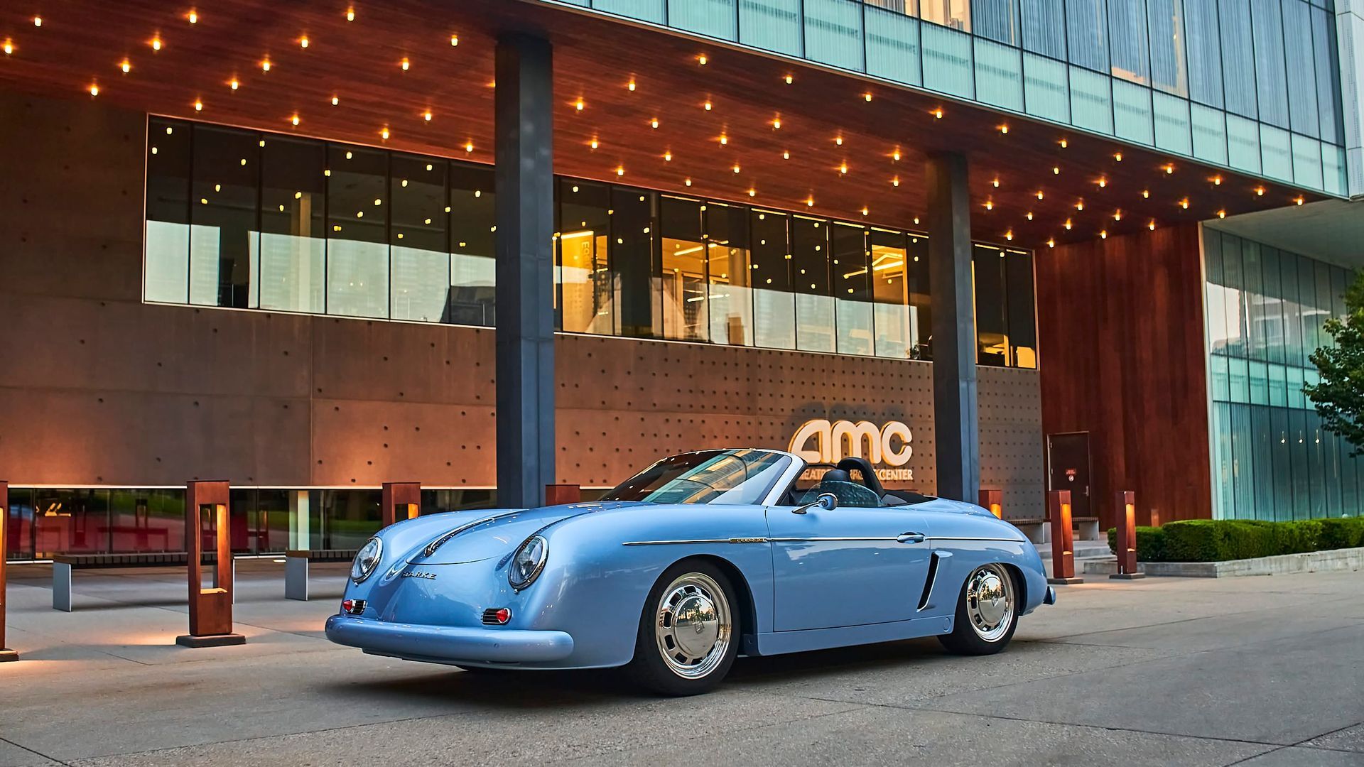 Light blue classic convertible parked in front of an AMC movie theater entrance. The building has a modern design.