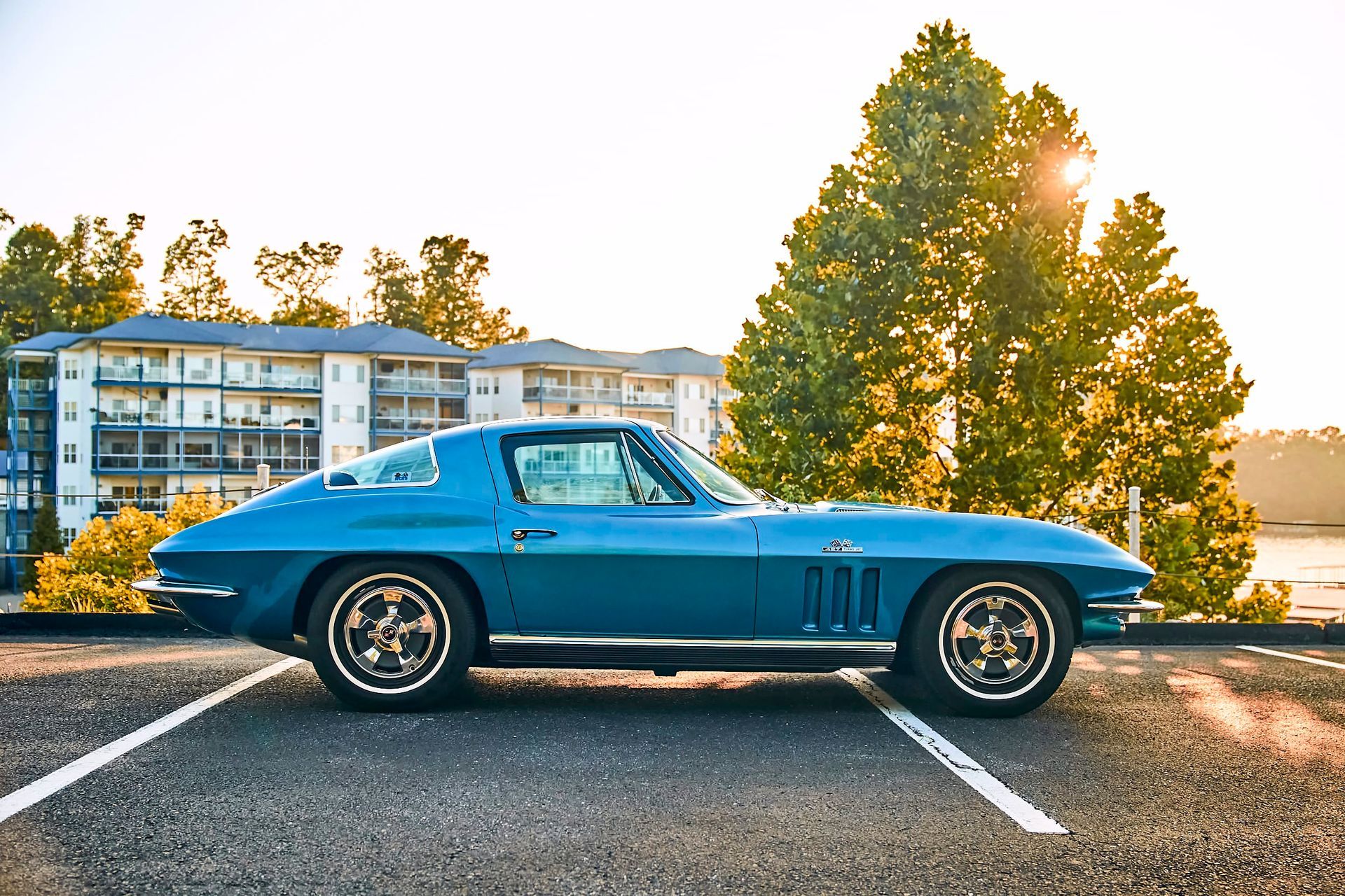 Blue classic Corvette parked in a parking lot with buildings and a tree in the background, illuminated by the sun.