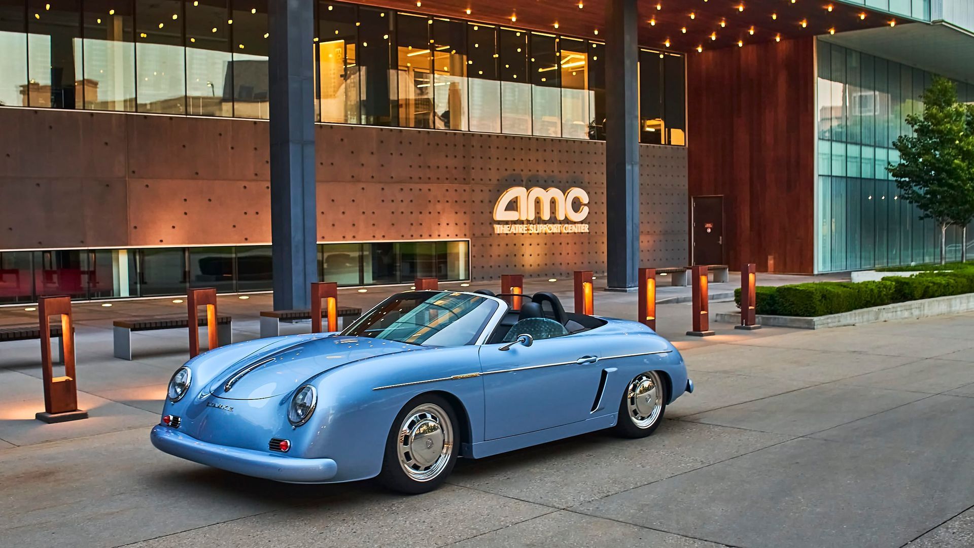 A light blue convertible parked outside an AMC theater. The car has a vintage design. The setting is modern.