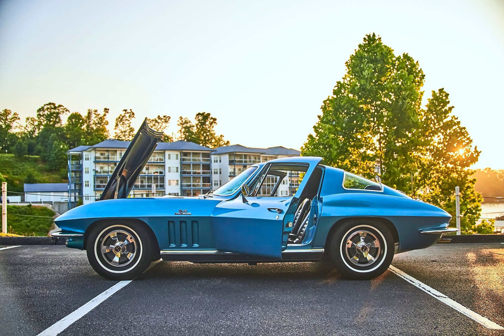 A classic blue Corvette coupe with the hood and door open, parked in front of a building and tree, with the sun setting in the background.