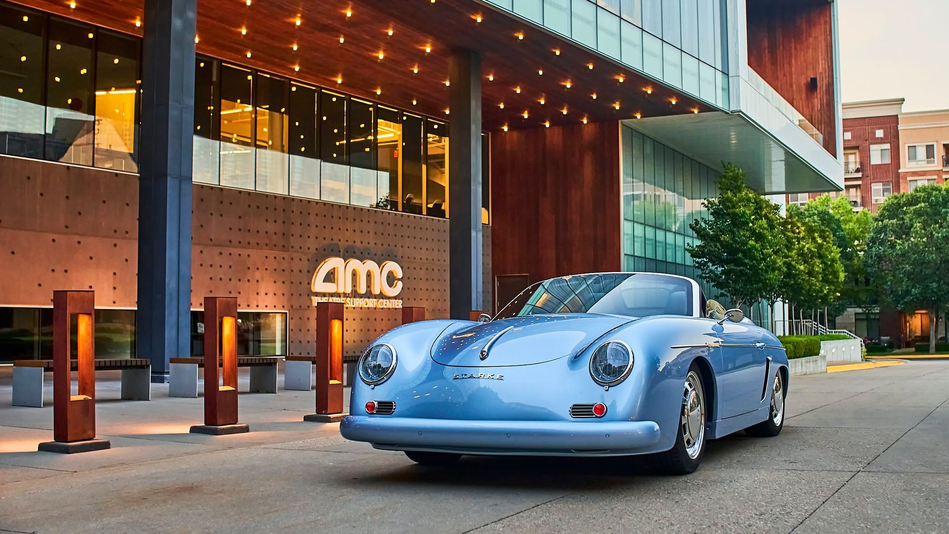 A light blue vintage car parked in front of an AMC theater. The building has modern architecture and is lit by warm lights.