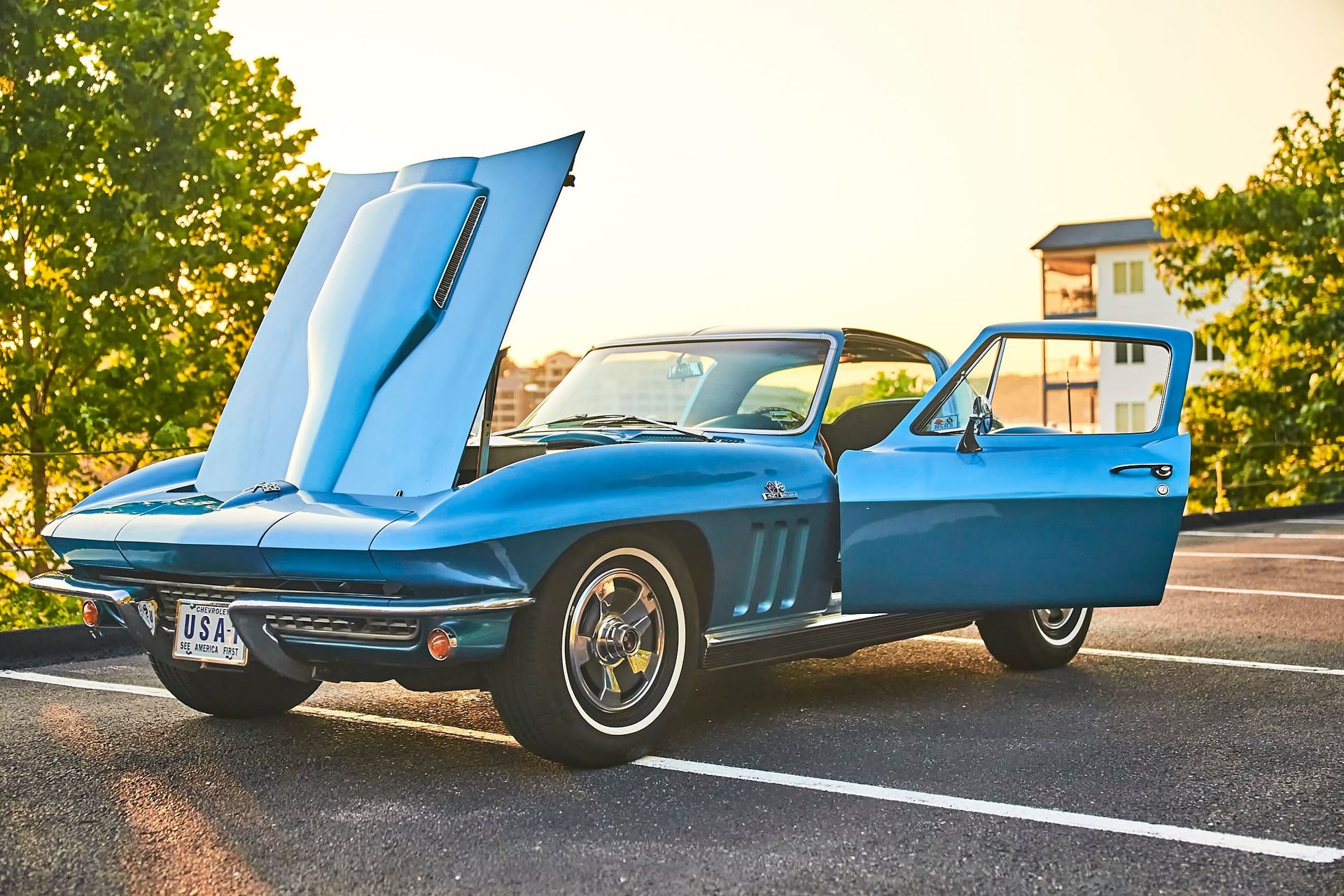 A blue vintage Corvette with its hood and door open parked on a paved surface, in front of trees.