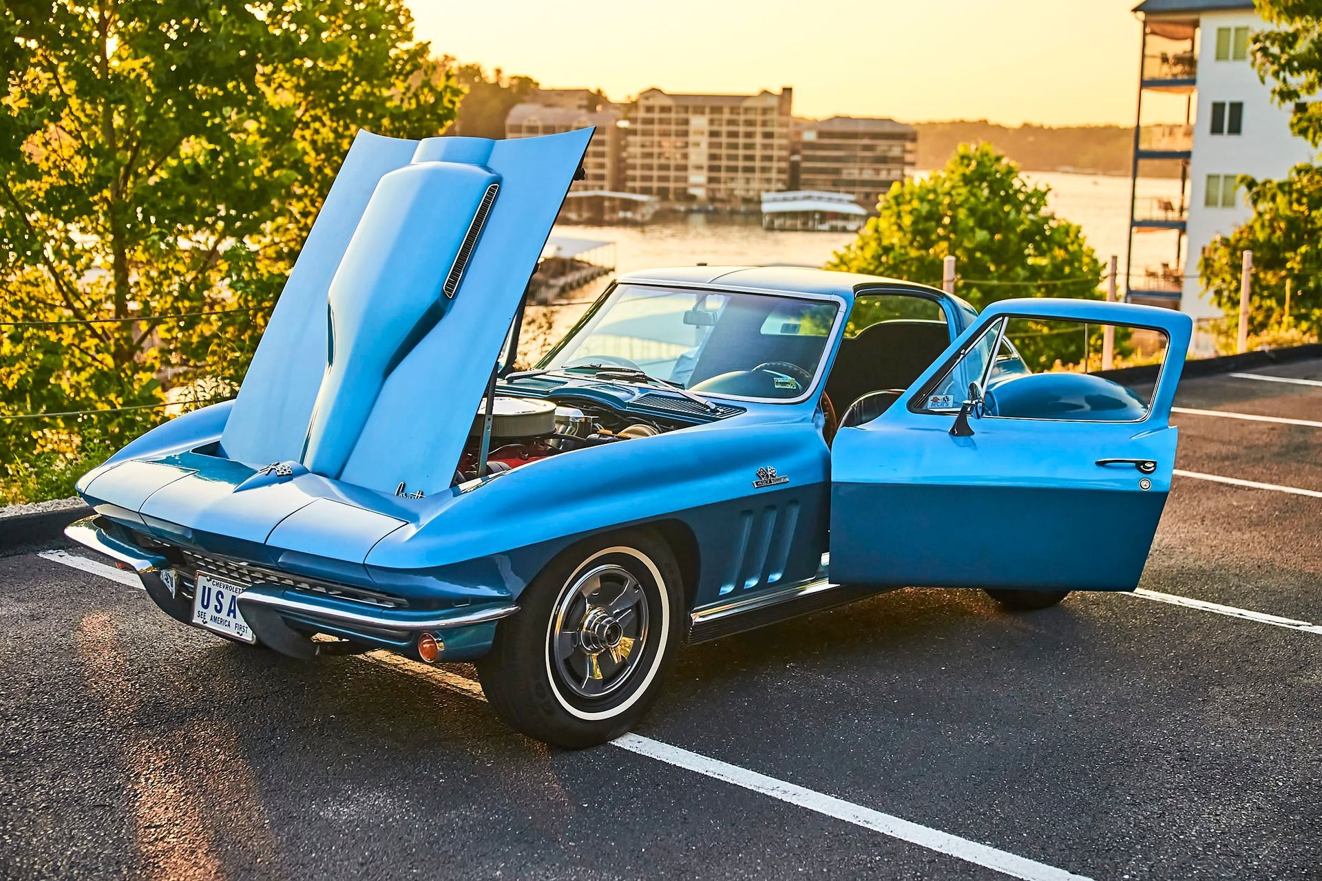 Blue vintage Corvette with hood and door open, parked on asphalt with a lake and buildings in the background.