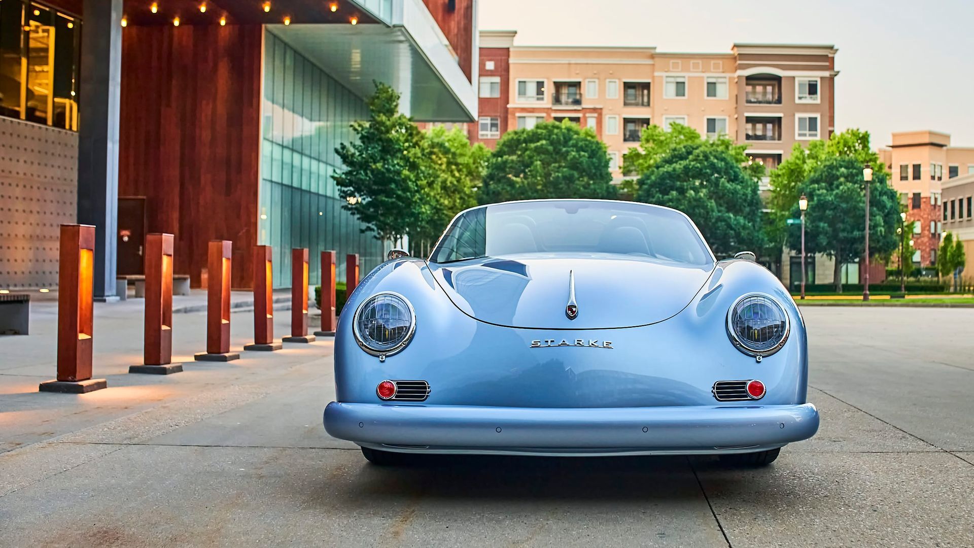 Pale blue vintage Porsche 356 parked in front of a modern building with wood accents and green trees in the background.