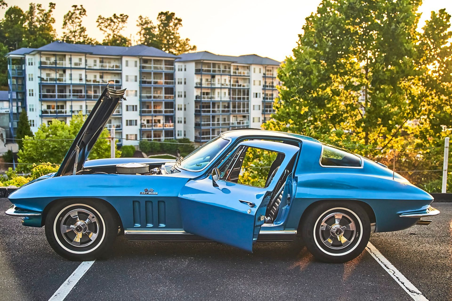 Blue vintage Corvette with hood and door open, parked in front of a multi-story building.