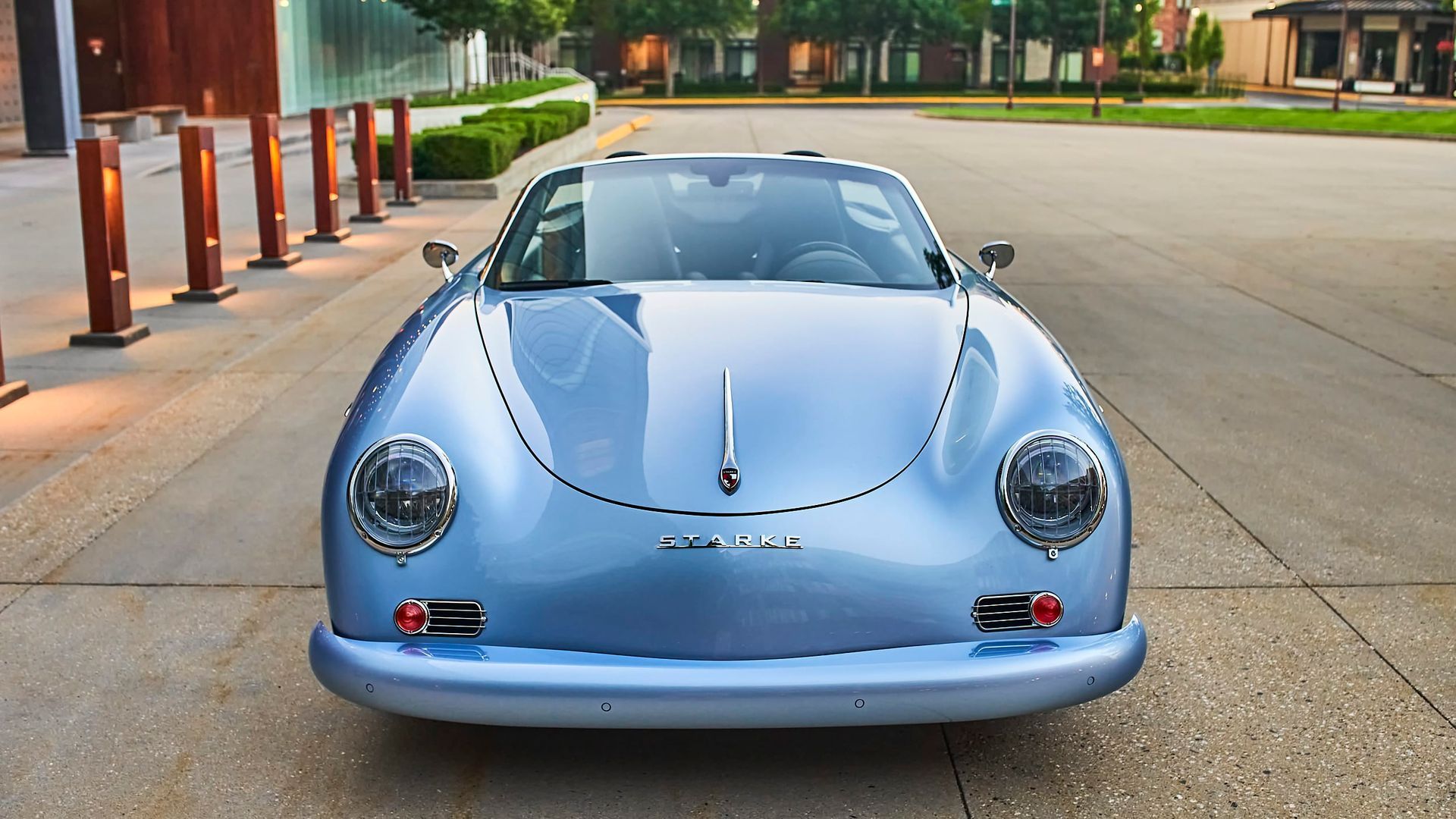Light blue vintage Porsche 356 convertible parked on a paved walkway. The car faces forward in a well-lit, urban setting.