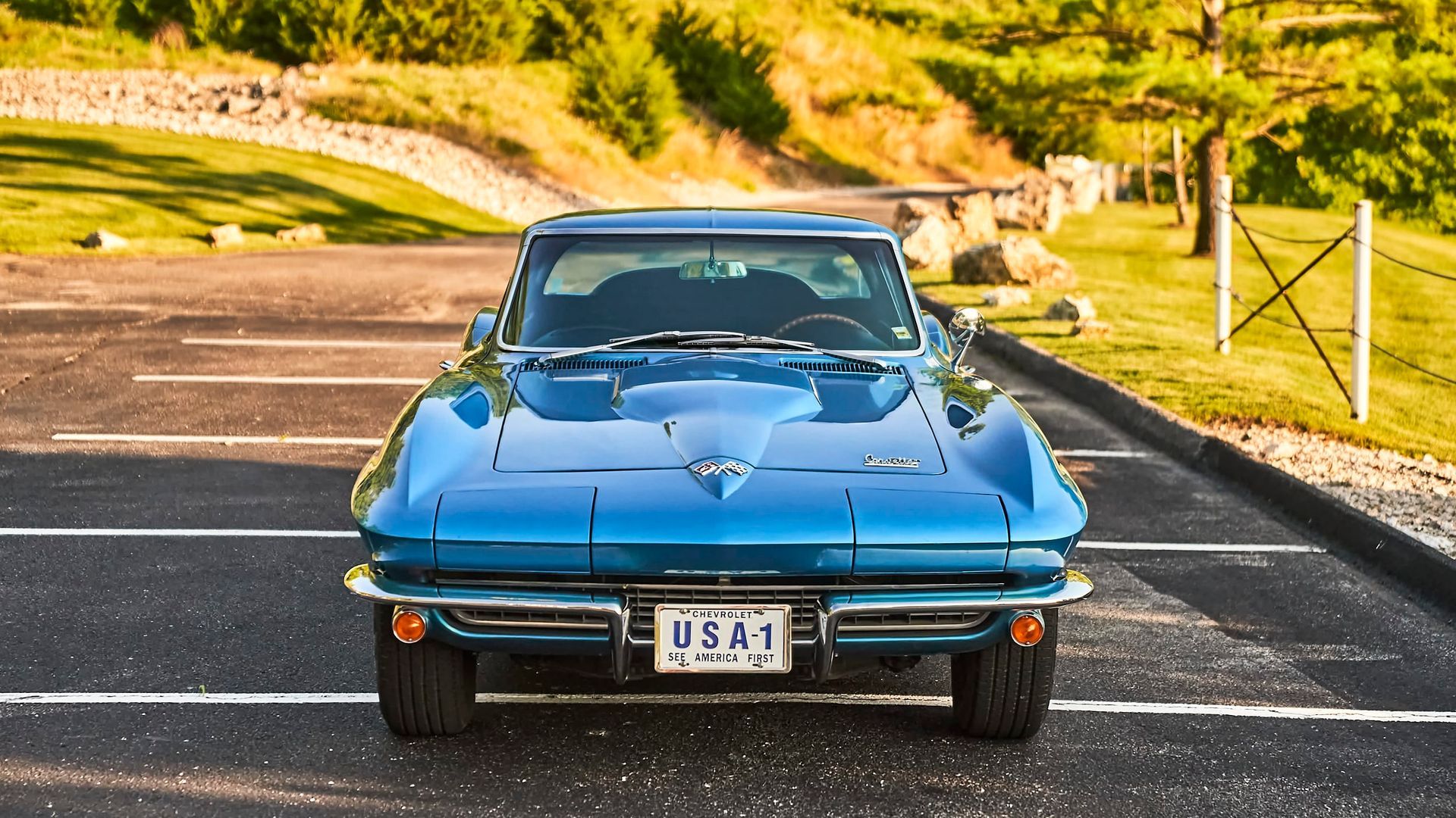Blue classic Corvette parked on asphalt, facing the viewer. The car is in a sunny outdoor setting.