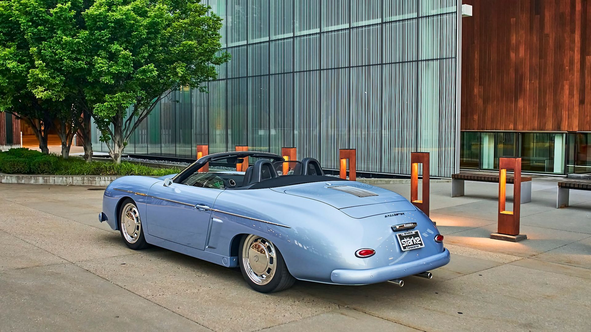 Pale blue classic convertible Porsche parked in front of a modern building with brown and gray panels; a tree is visible to the left.