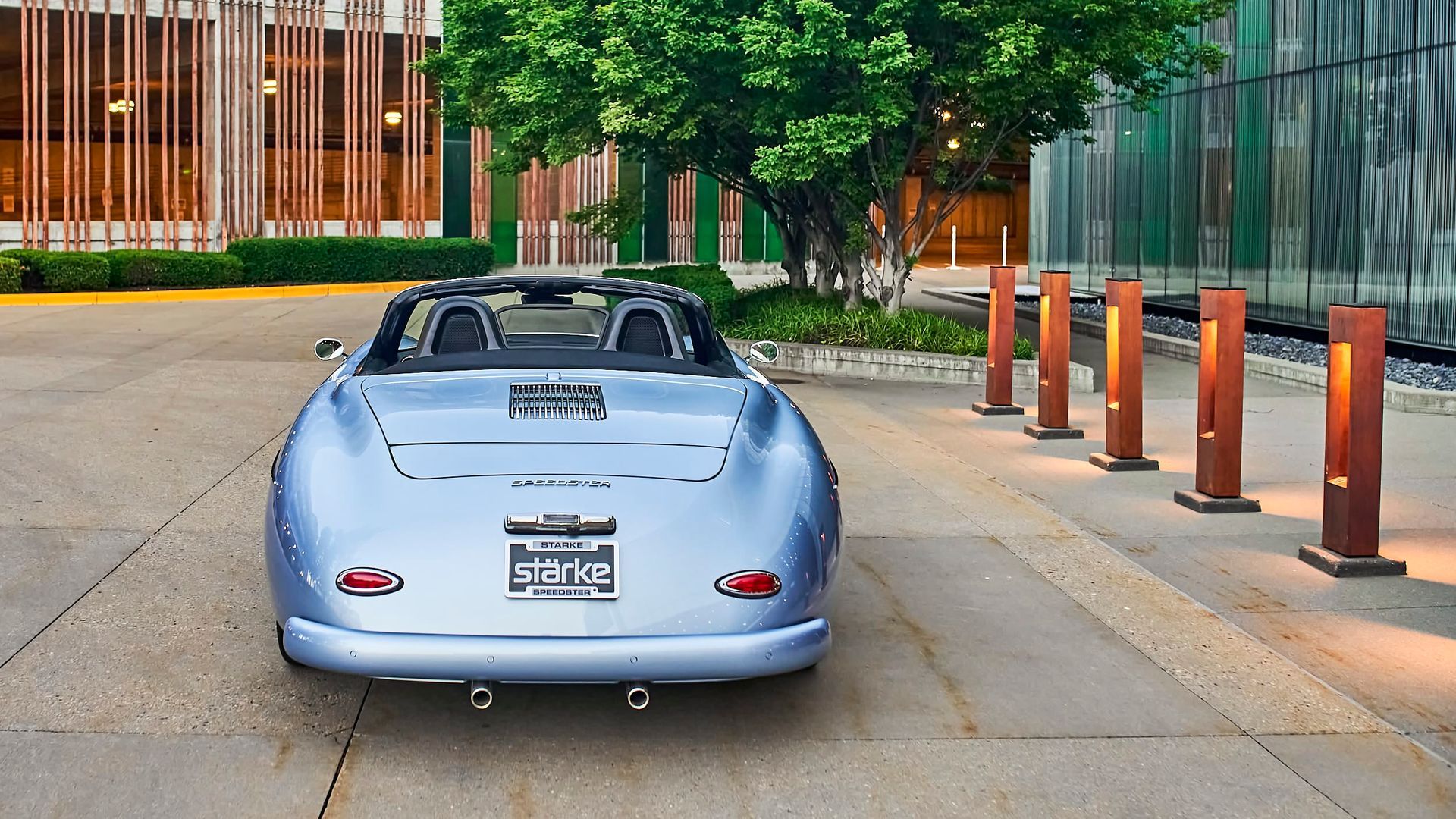 A light blue vintage Porsche convertible parked outside a modern building with brown pillars and a tree.