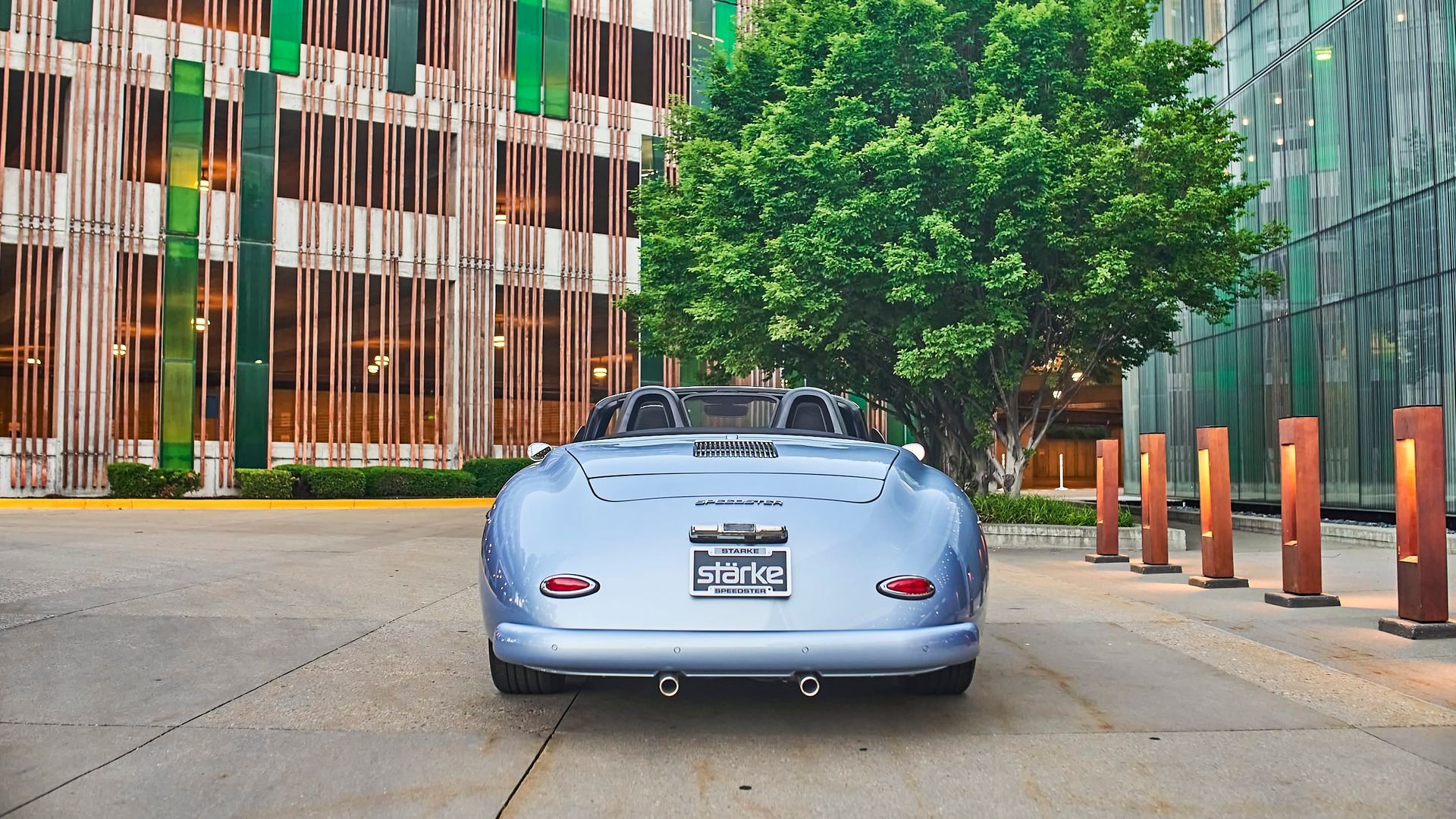 Rear view of a light blue vintage convertible parked in front of a modern building with a small tree.