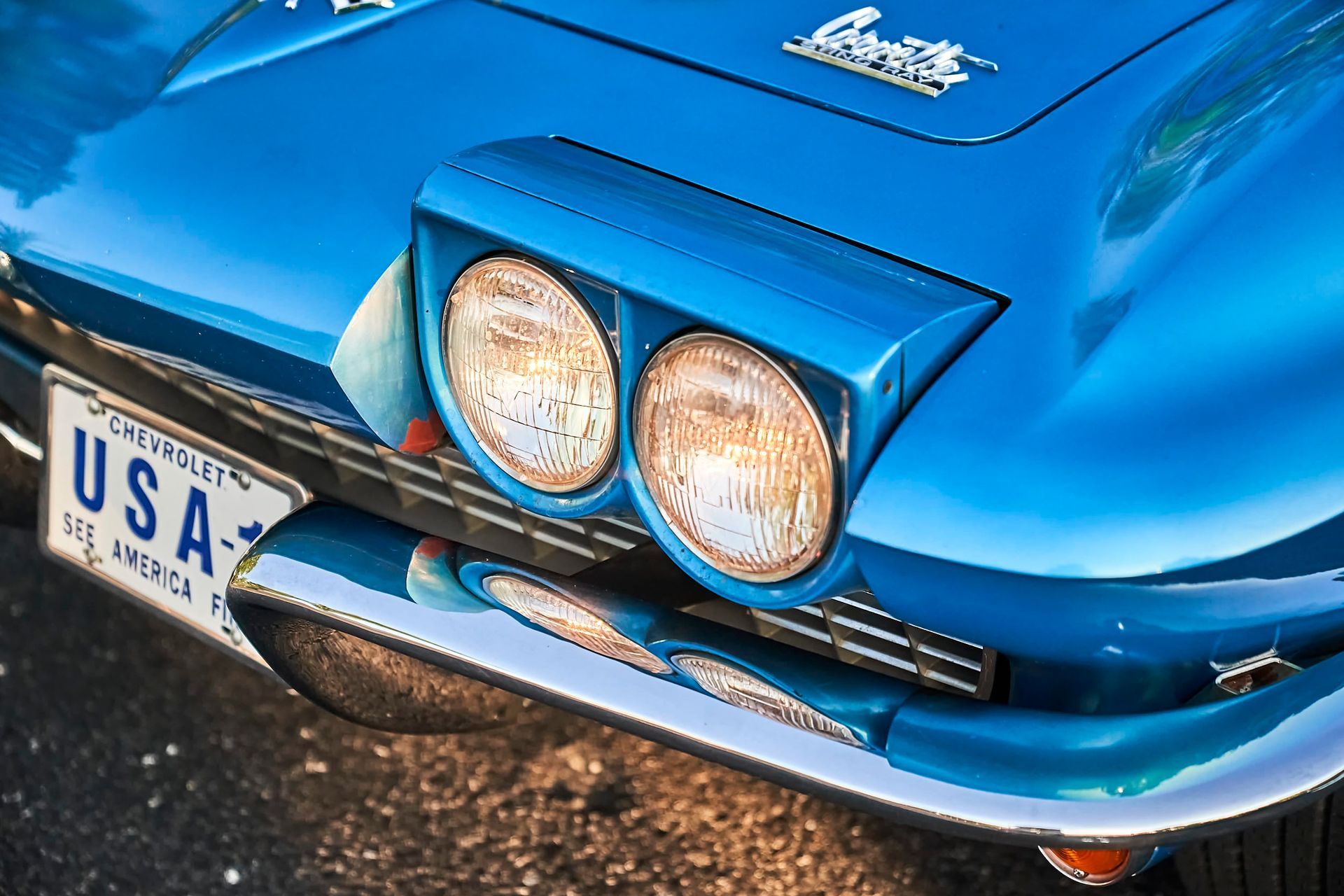 Close-up of a classic blue Corvette with chrome accents and exposed headlights, displaying a USA license plate.