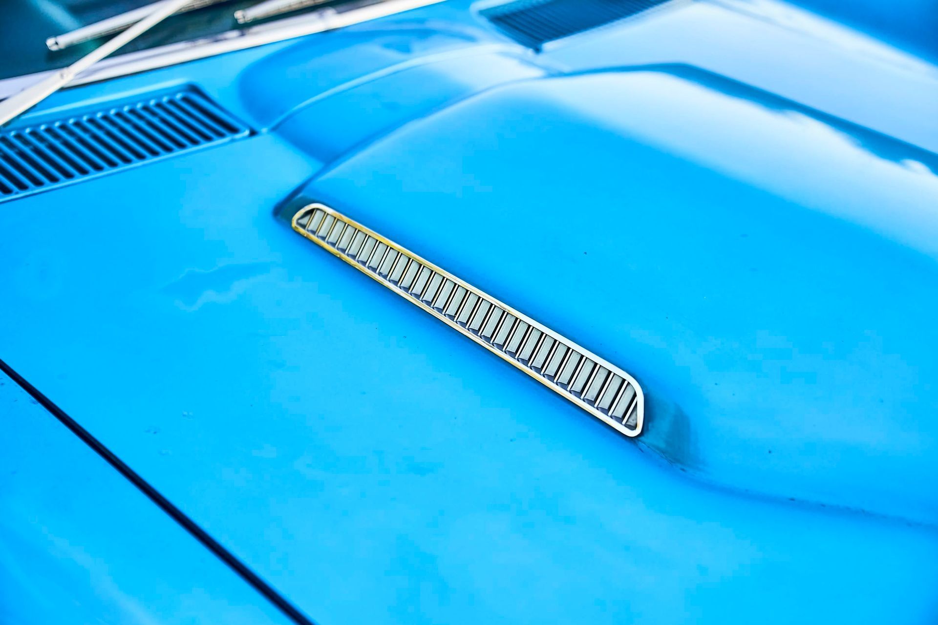 Close-up of a bright blue car hood with a silver air vent. Windshield wipers are visible in the background.