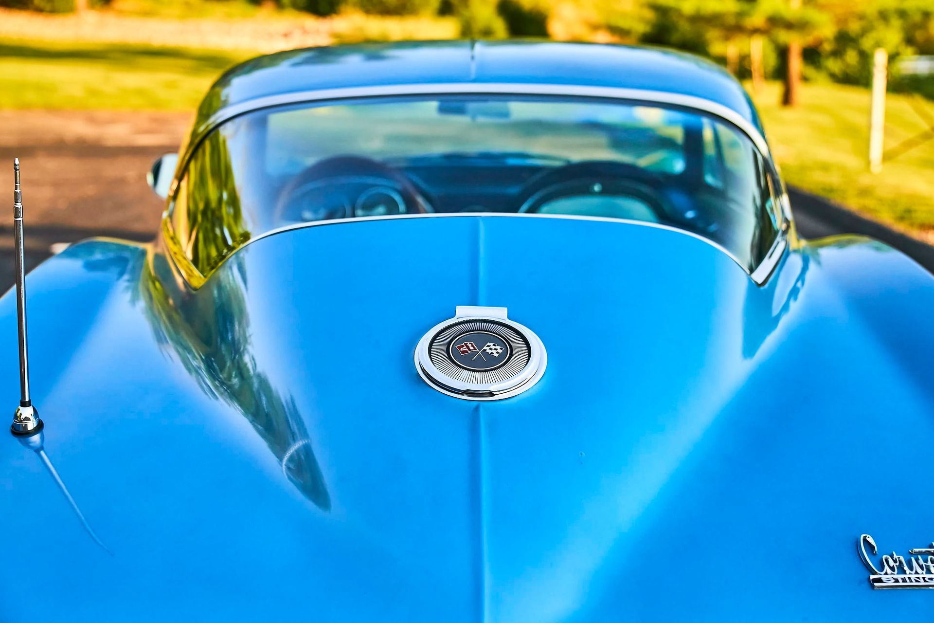 Blue vintage Corvette, close-up of hood with emblem. Windshield reveals steering wheel. Antenna on the left.