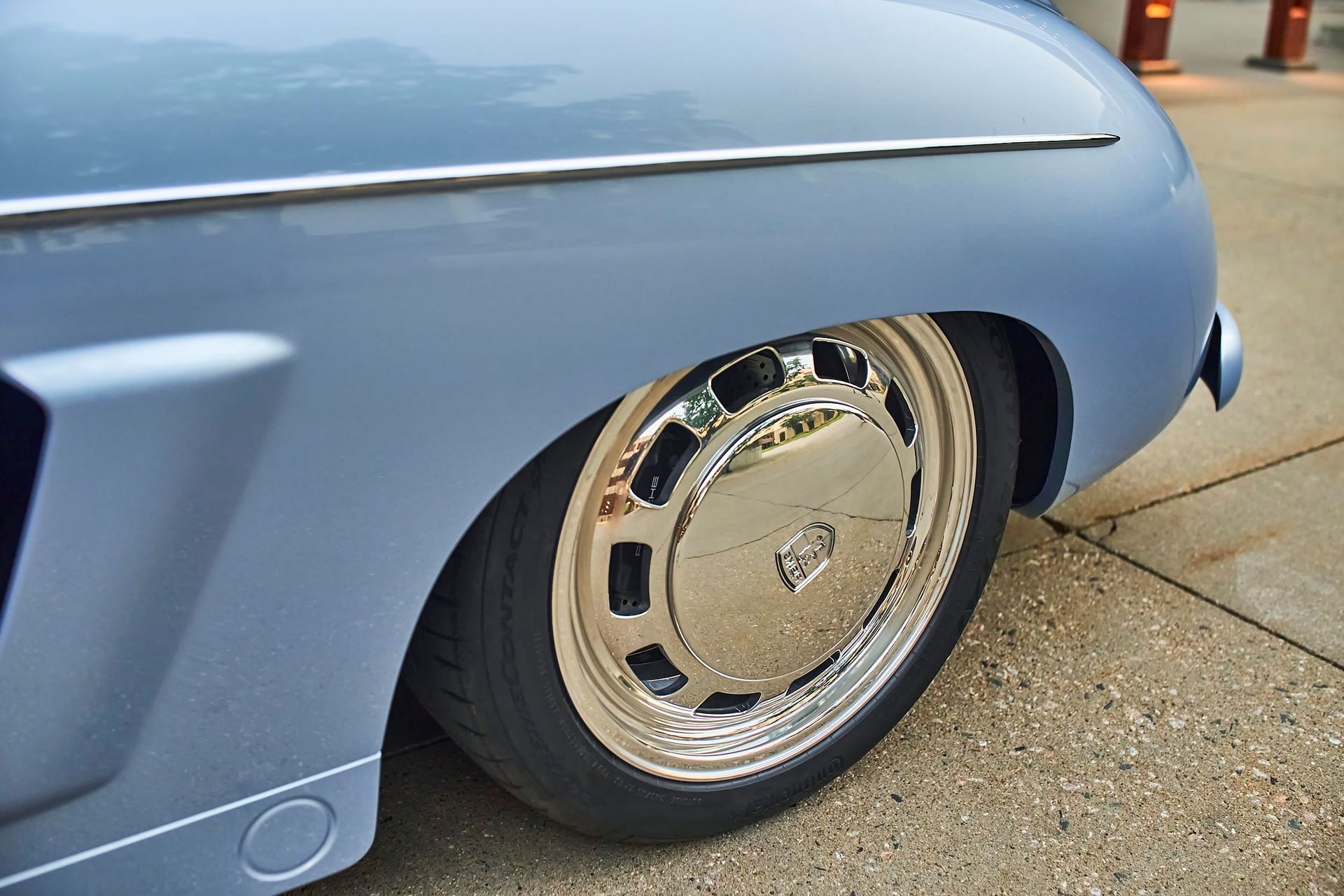 Close-up of a light blue vintage car's front wheel with a chrome hubcap, set against a concrete background.