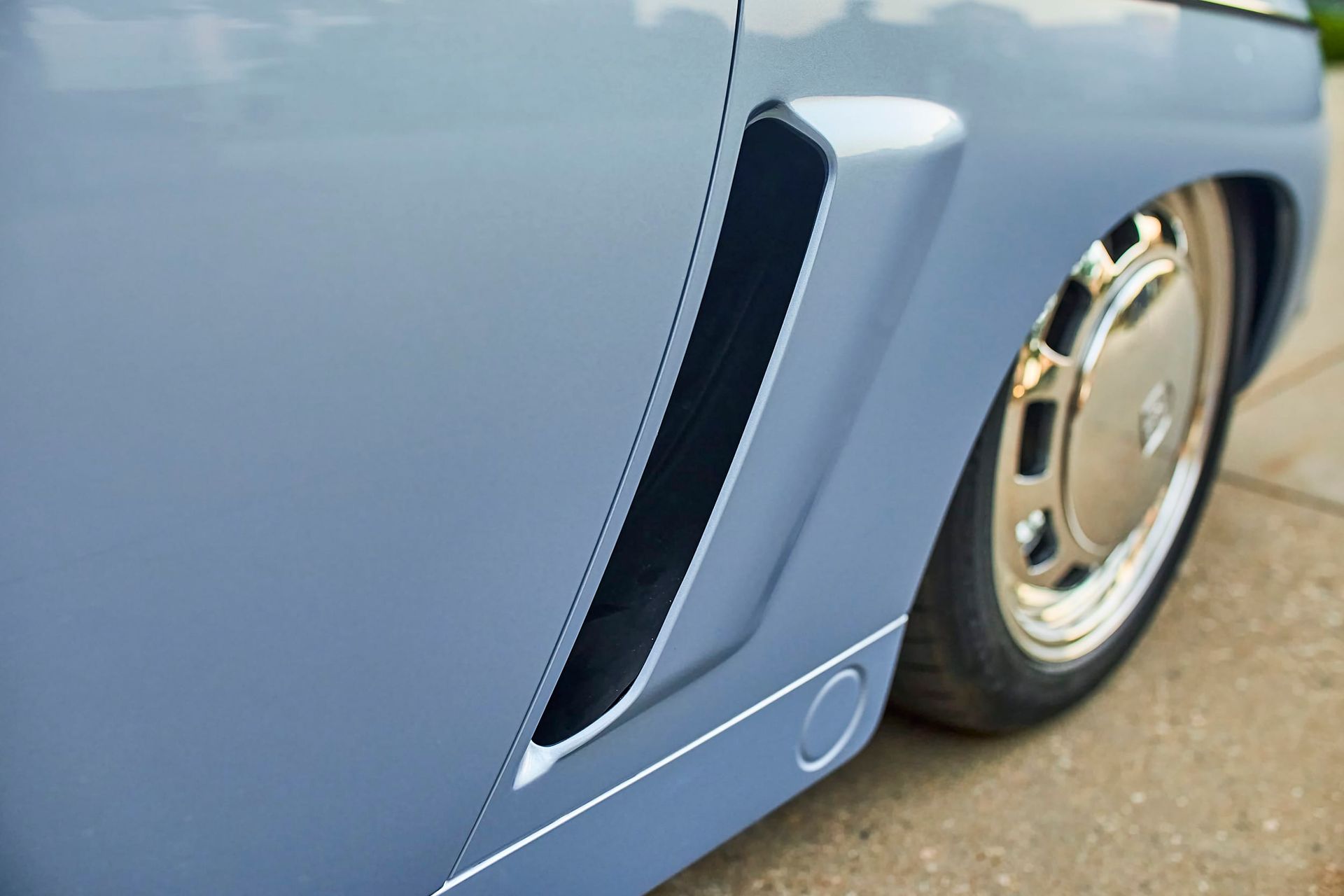 Close-up of a gray car's side panel with a black vent, and a chrome wheel.
