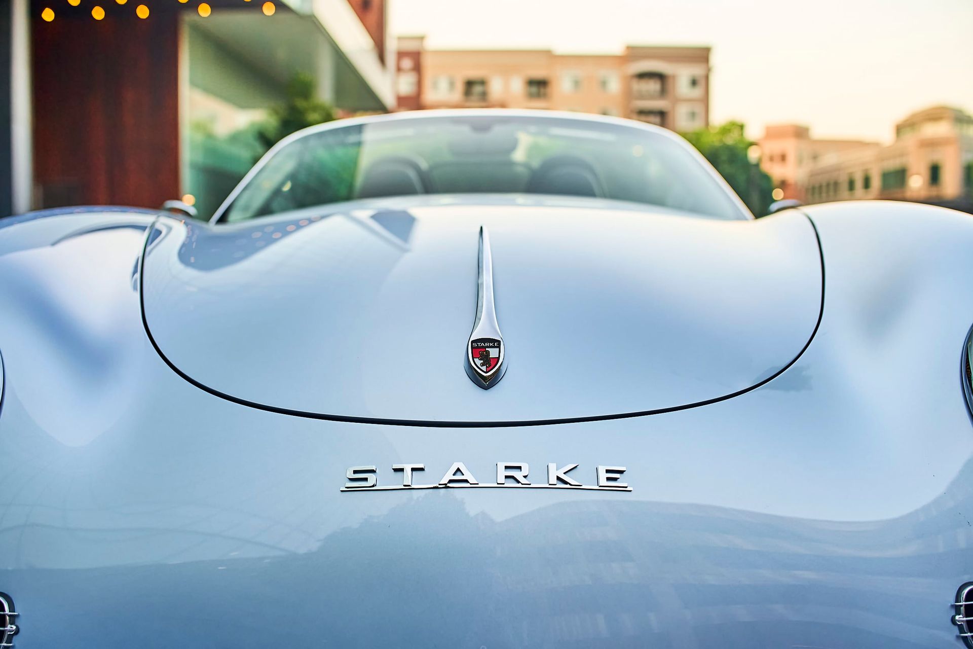 Close-up of a light blue Starke roadster car with the hood up, parked outdoors in a sunny setting.
