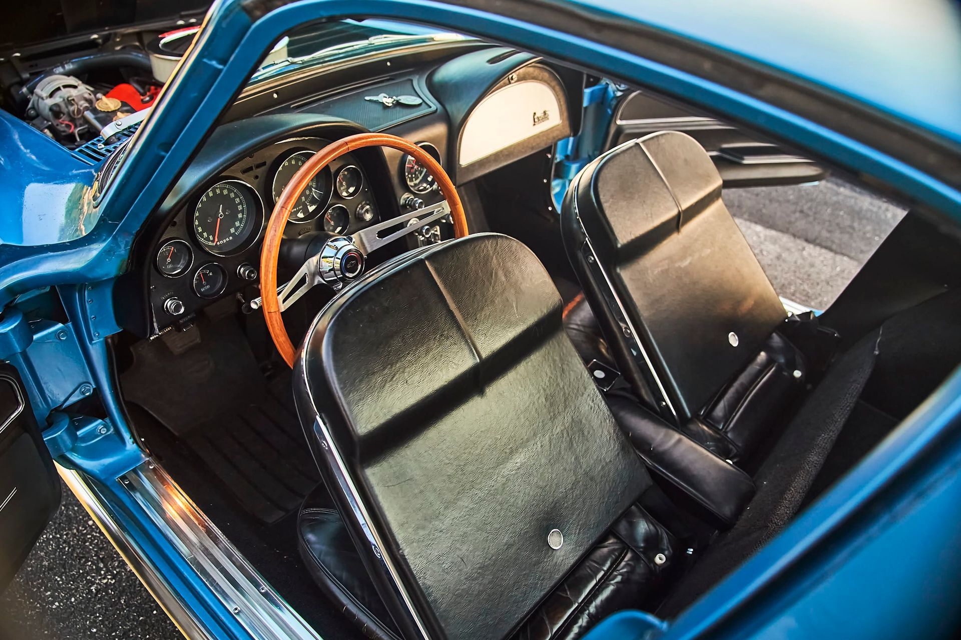 Interior of a classic blue Corvette. Black leather seats, wooden steering wheel, and vintage gauges are visible.