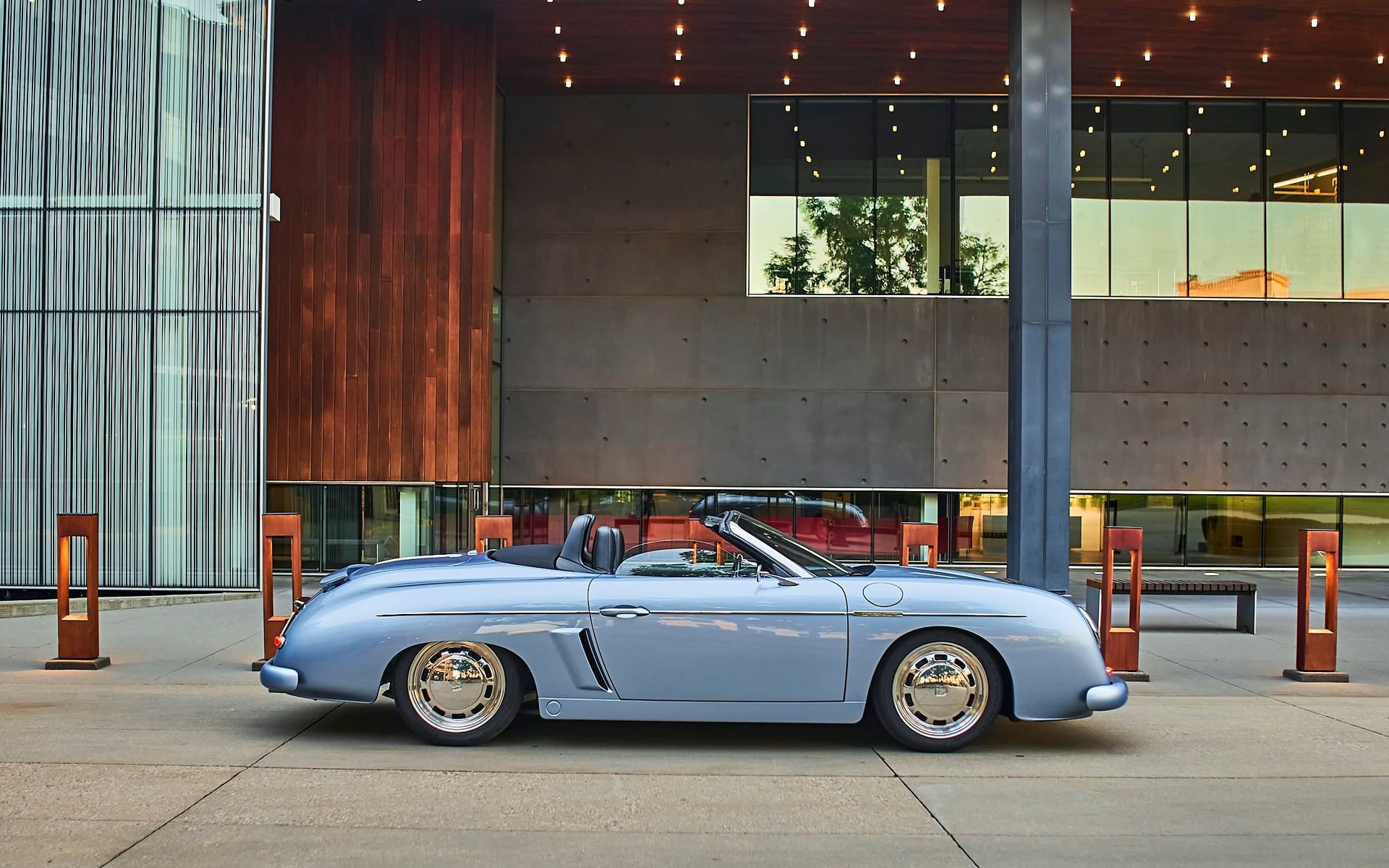Light blue classic convertible parked in front of a modern building with a reflective glass facade and wooden accents.