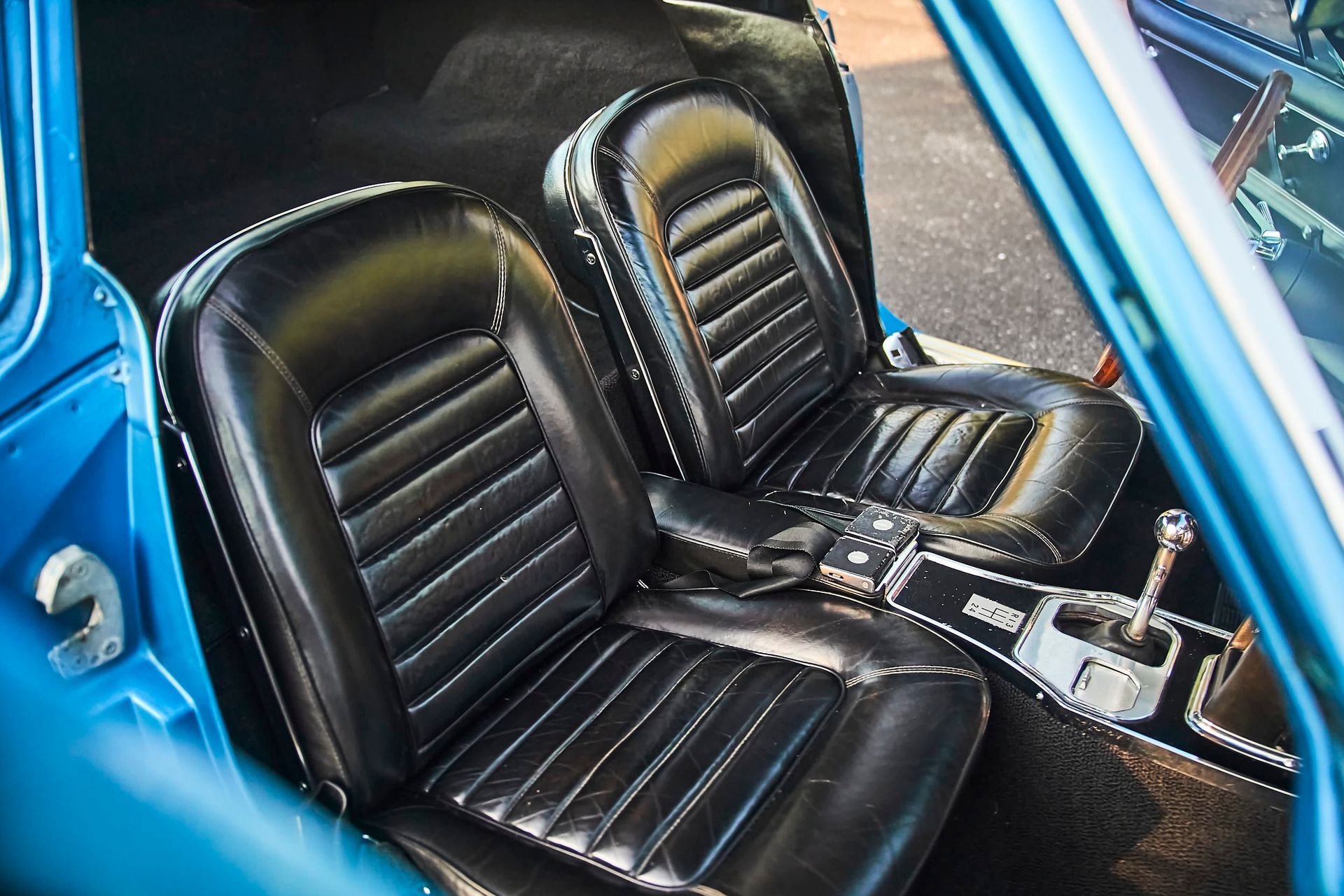 Black leather car seats with ribbed detailing, set in a blue car interior. The seats face forward, with a center console visible between them.