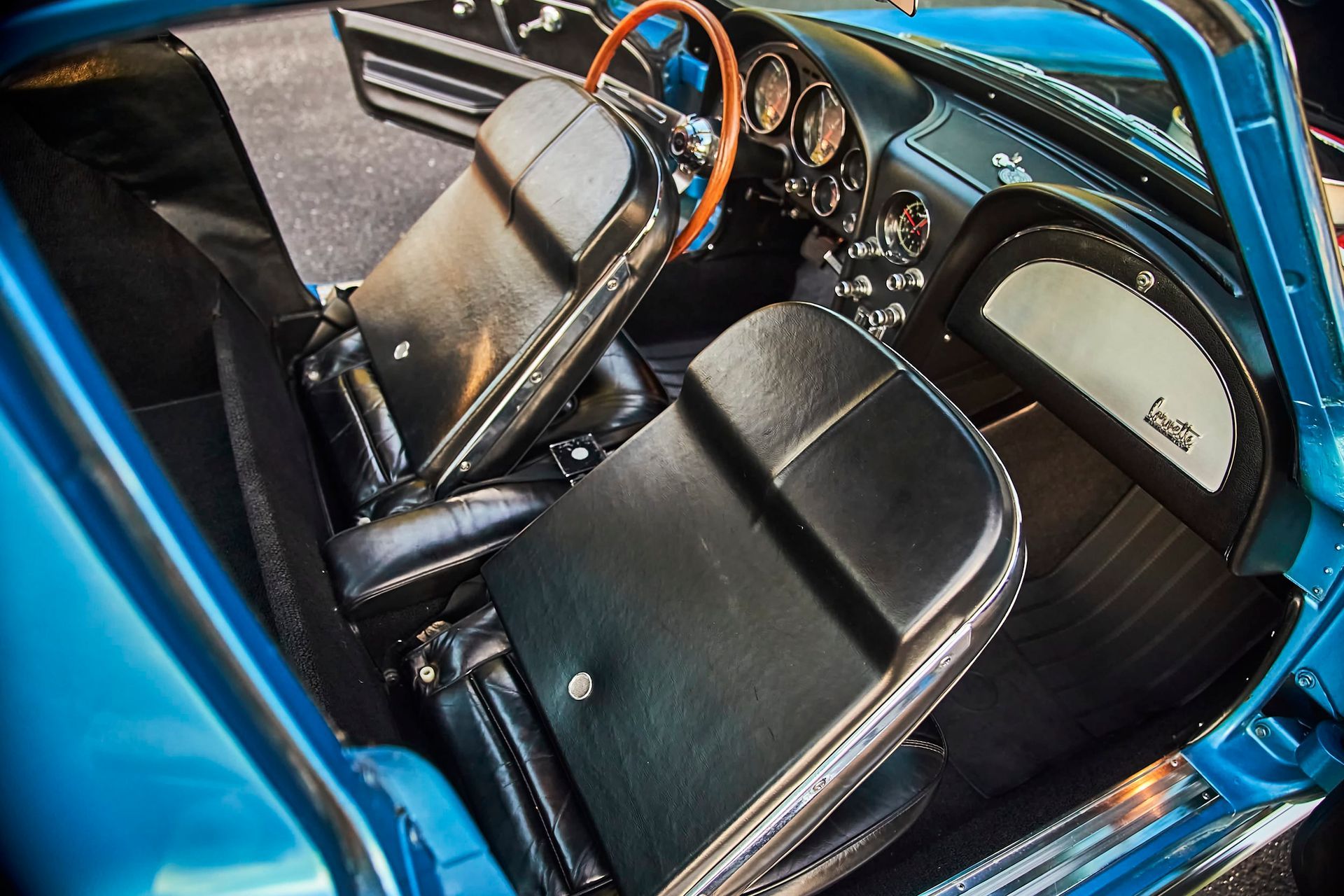 Interior of a classic blue Corvette, showing black leather seats, wooden steering wheel, and dashboard.