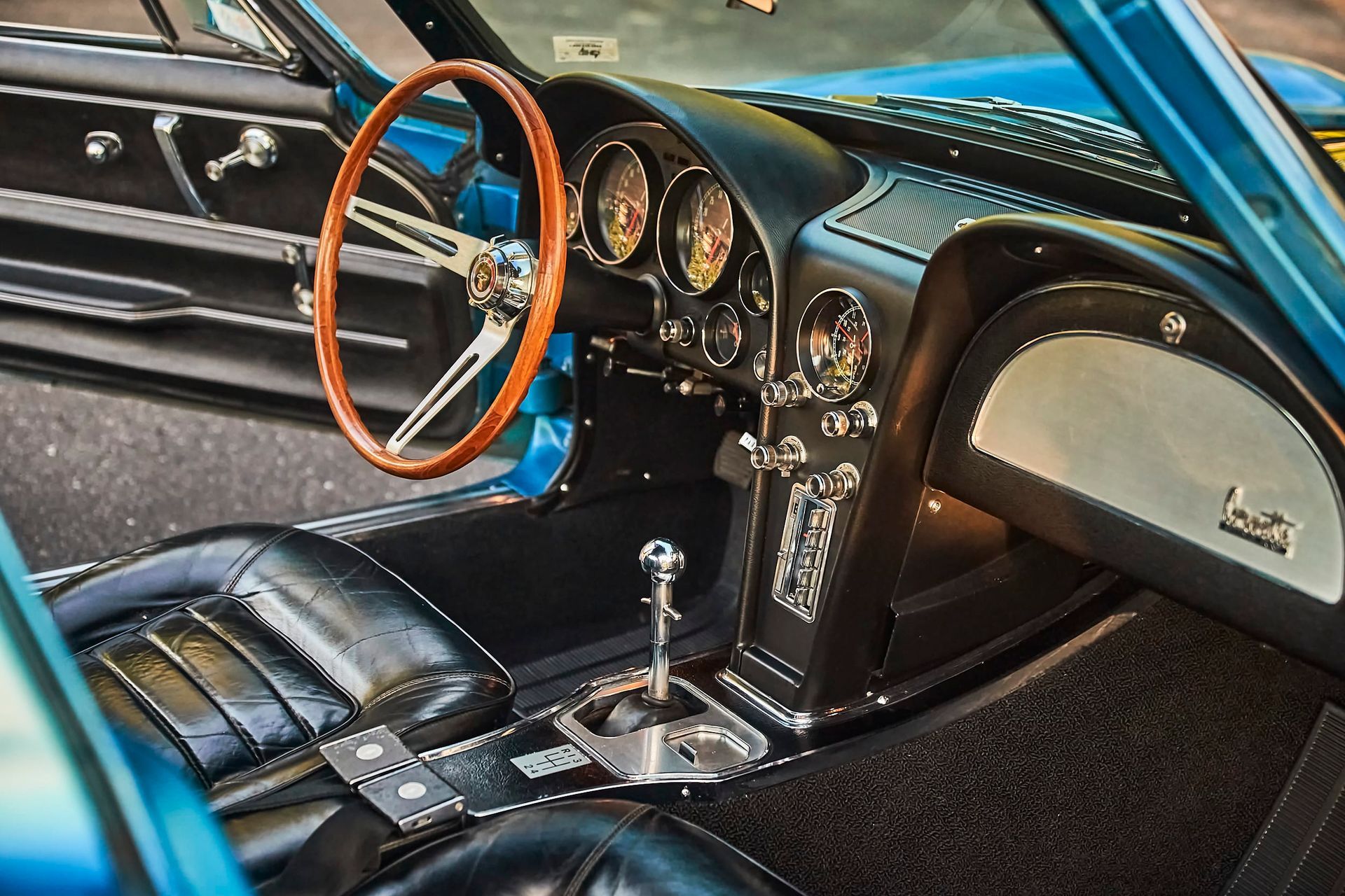 Interior of a classic blue Corvette. Features include a wood steering wheel, black leather seats, chrome detailing, and a manual transmission.