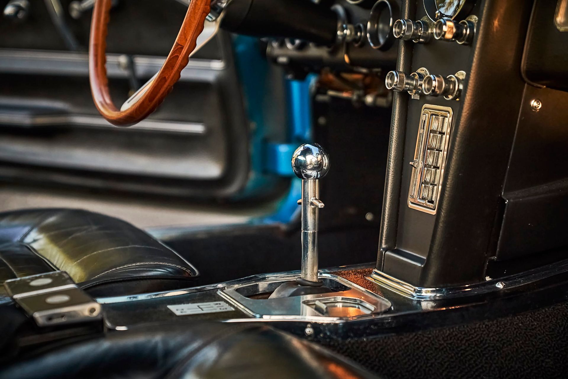 Interior of a classic car. Focus on a chrome gear shift, wooden steering wheel, and black leather seats.