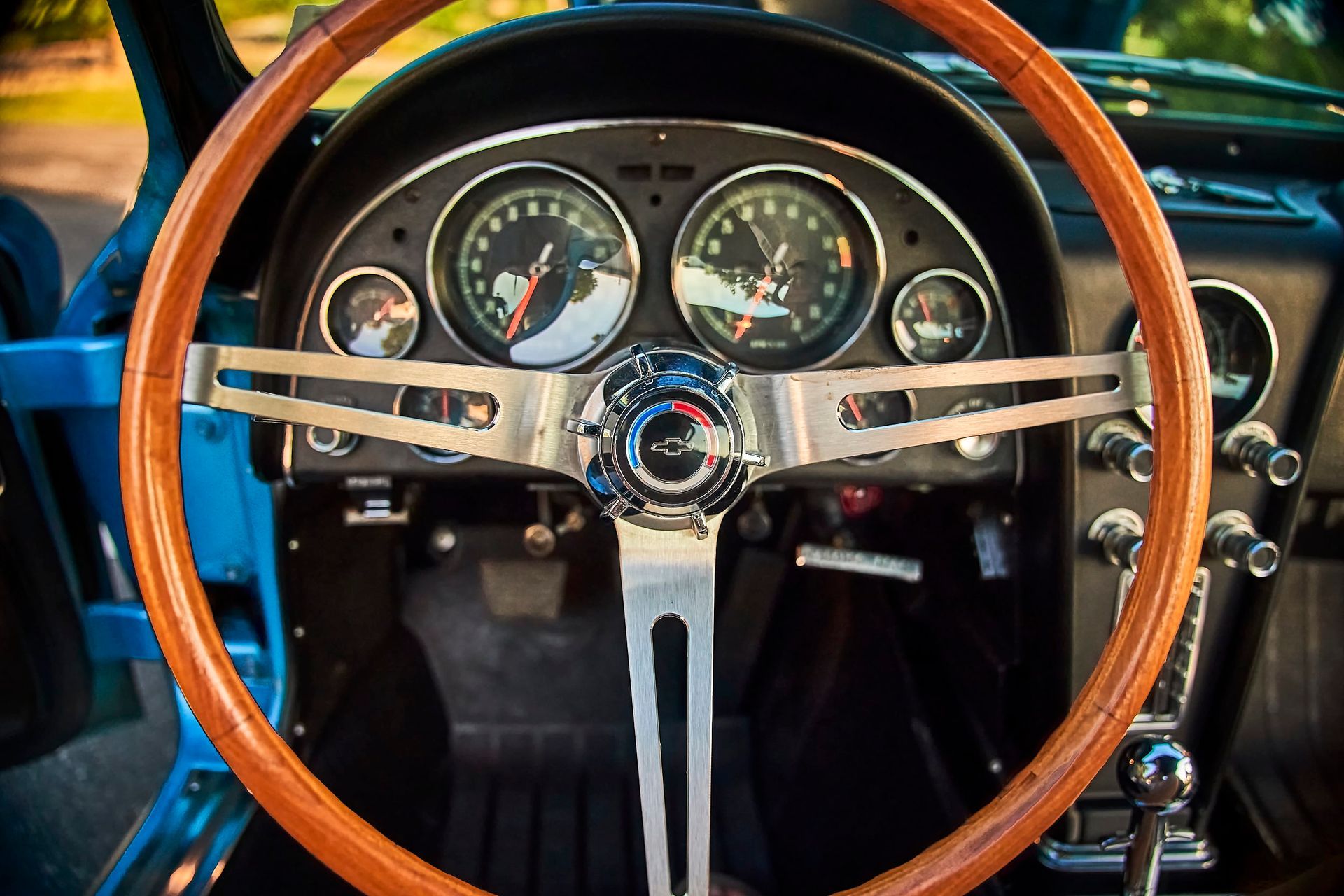 Classic car dashboard with wooden steering wheel, chrome accents, and round gauges in a blue interior.