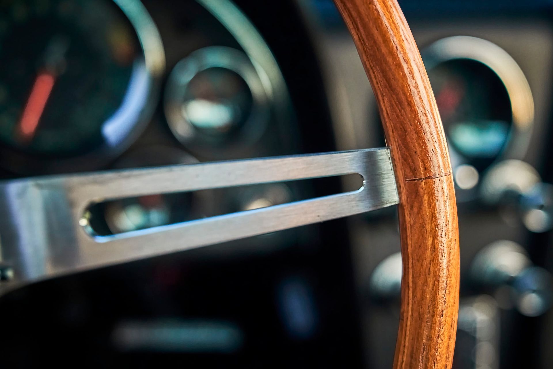 Close-up of a classic car's steering wheel with a wood-grain rim and silver spokes, set against the blurred dashboard gauges.
