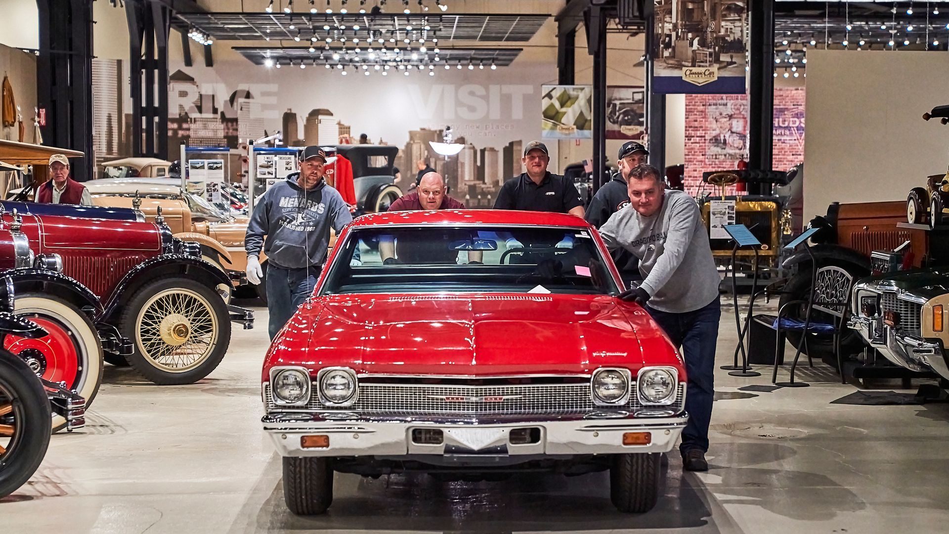 A group of men are standing around a red car in a museum.