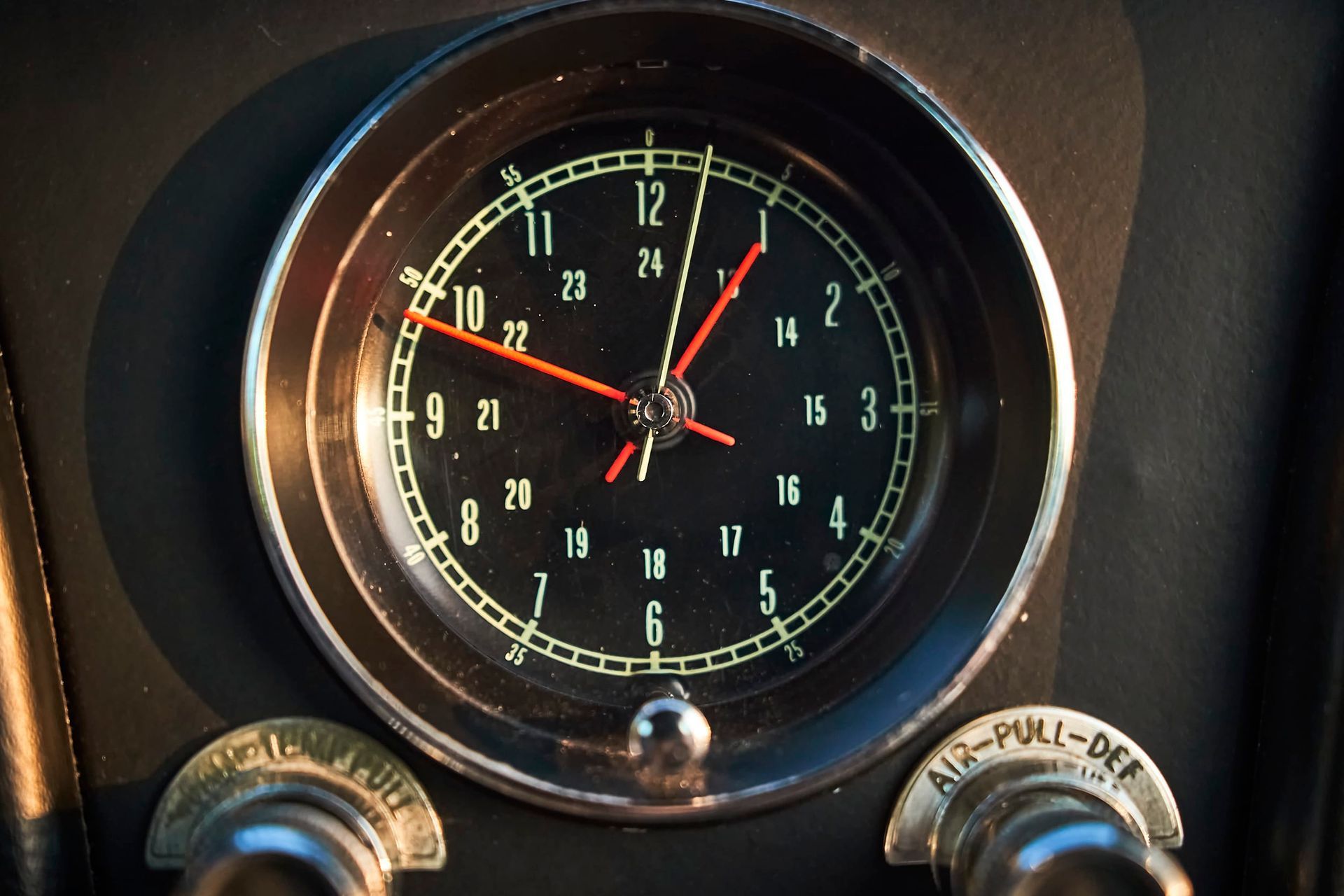 Close-up of a car clock with a black face and red hands, surrounded by a chrome bezel, with the text 