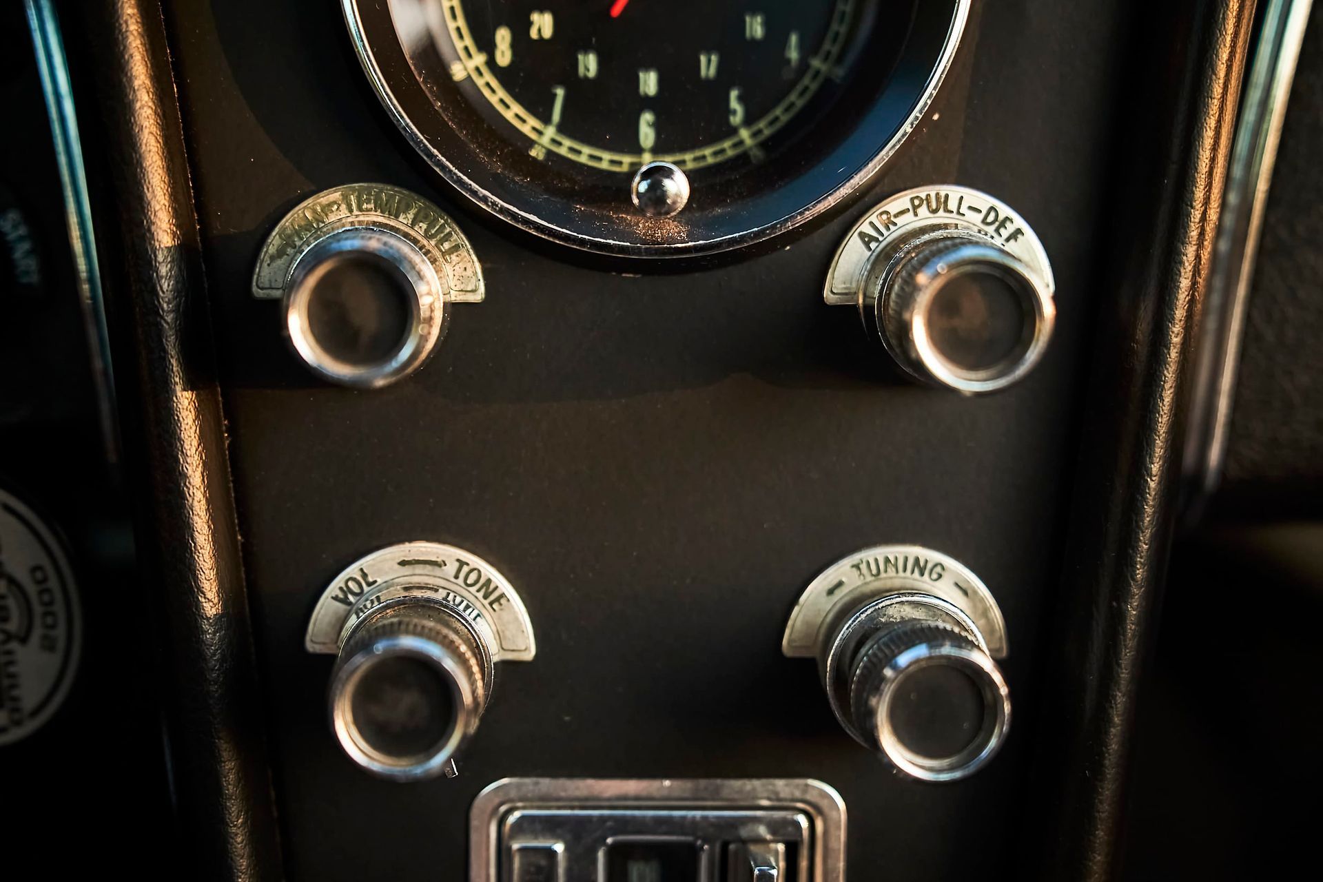 Close-up of a classic car dashboard with knobs for fan/heat, radio, and more; clock visible.