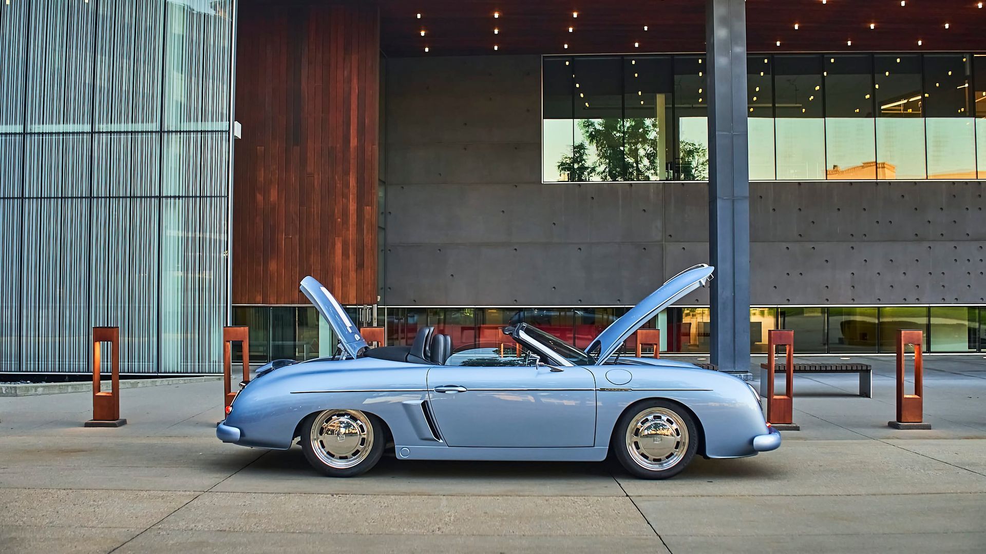 Light blue vintage convertible with open hoods parked outside a modern building with wood and glass accents.