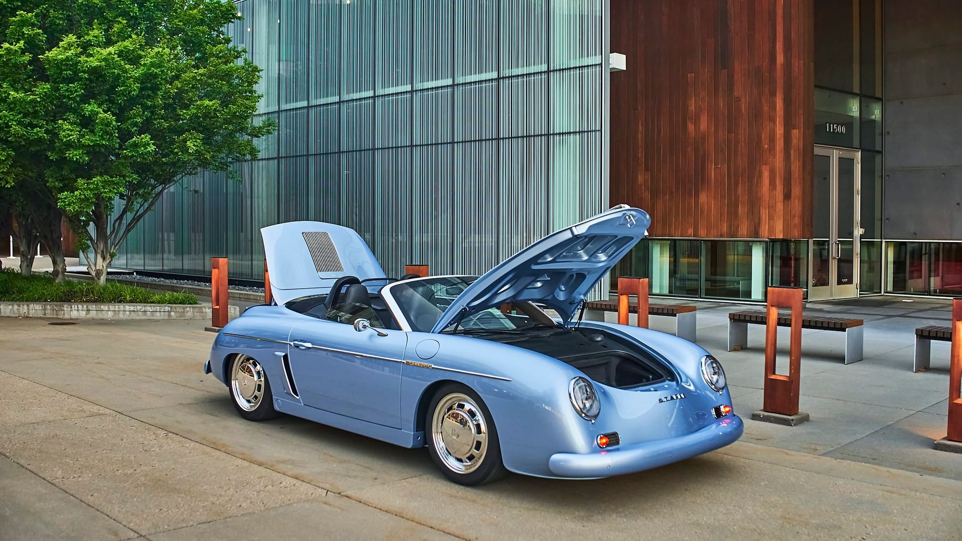 Light blue Porsche 356 Speedster convertible with an open hood, parked in front of a modern building with a tree.