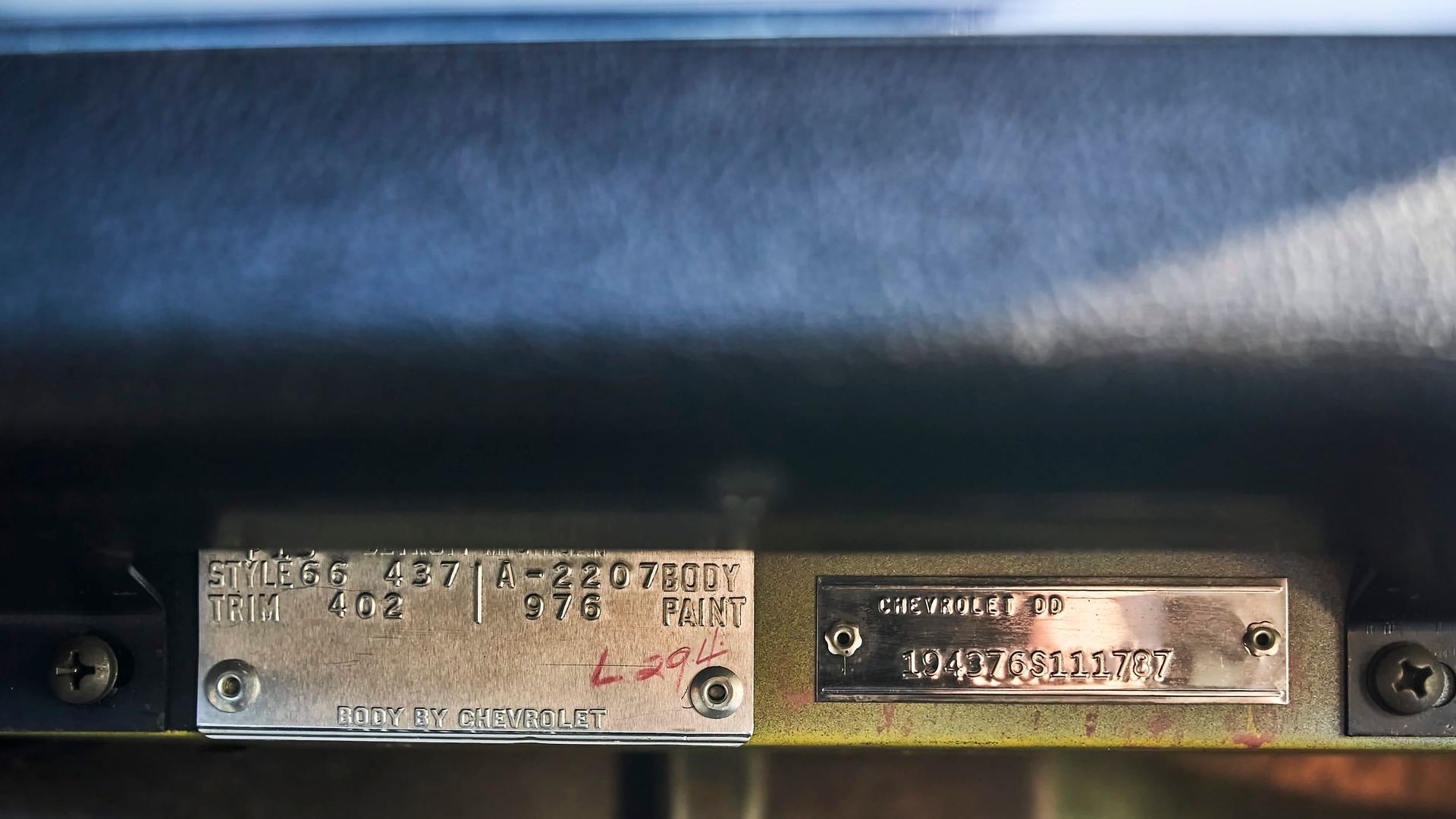 Two metal plates with stamped text, likely vehicle identification, attached to a dark-colored surface. The left plate is partially obscured by red ink.