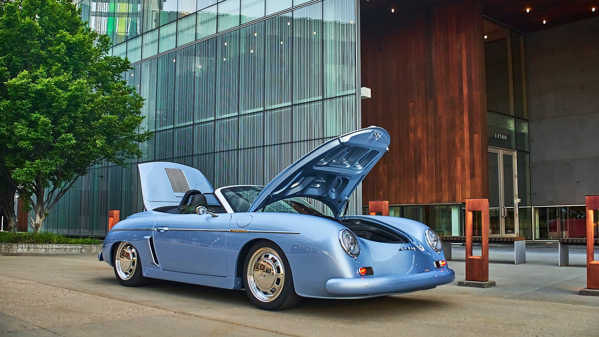 A light blue convertible sports car with its hood open, parked outside a modern building with a brown wood facade.