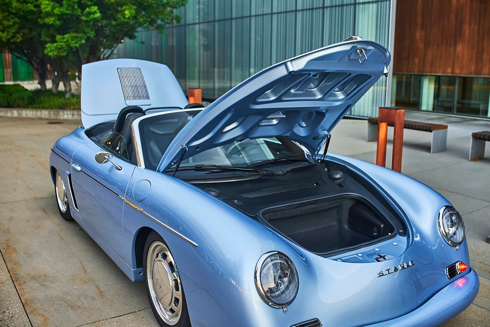 Light blue convertible Porsche with both the hood and trunk open, parked in a modern outdoor setting with glass and wooden structures.